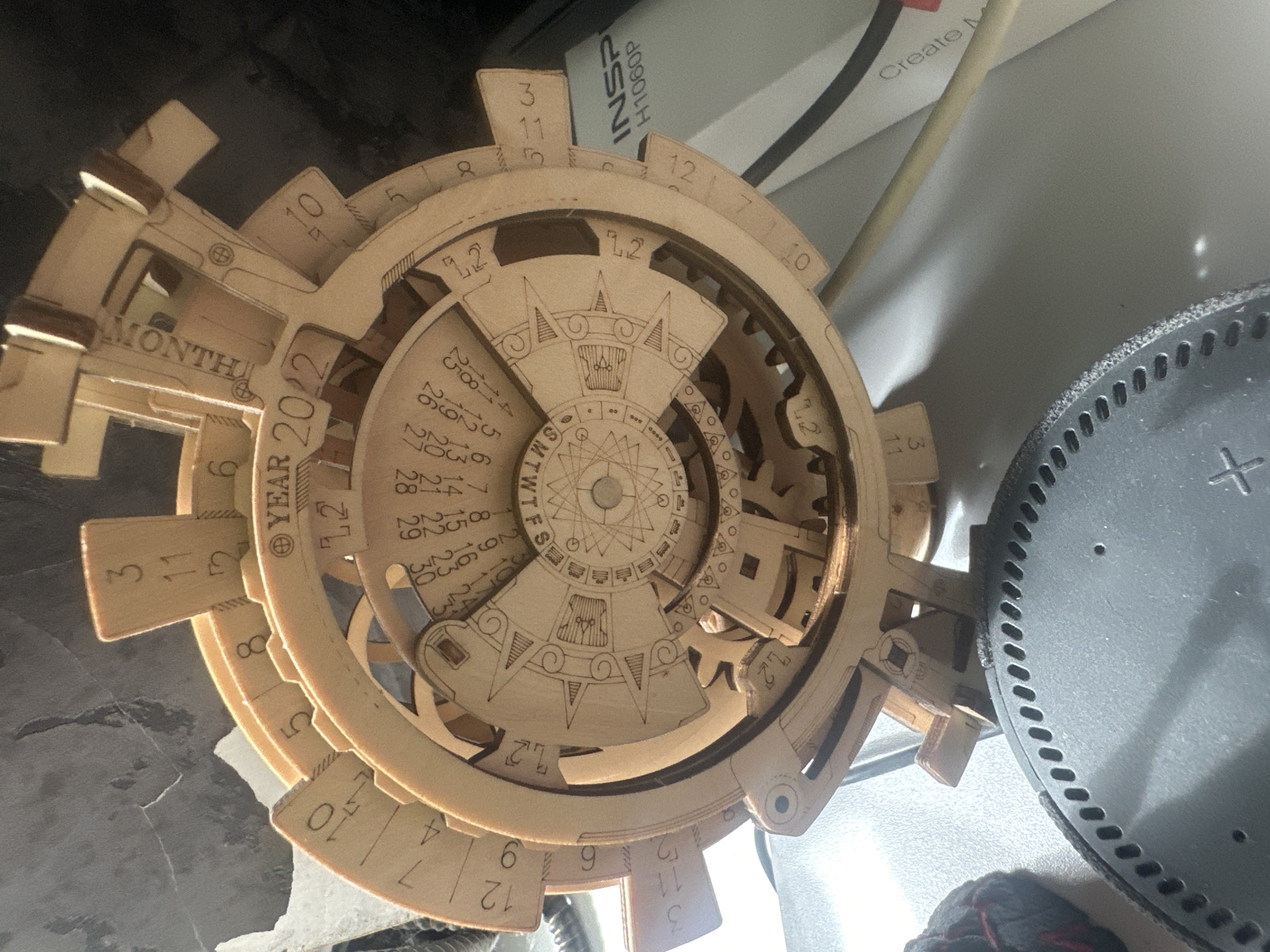 A wooden puzzle, likely a calendar or astrological device, is displayed on a white surface. The puzzle features intricate wooden gears and carved numbers and symbols. Partially visible in the background is a black circular object, possibly a speaker, and a white box with text that includes "Create M". The scene appears to be indoors, with a subtle shadow cast on the white surface. There are no people present in the image. The location is identified as Maladzyechna, Belarus.