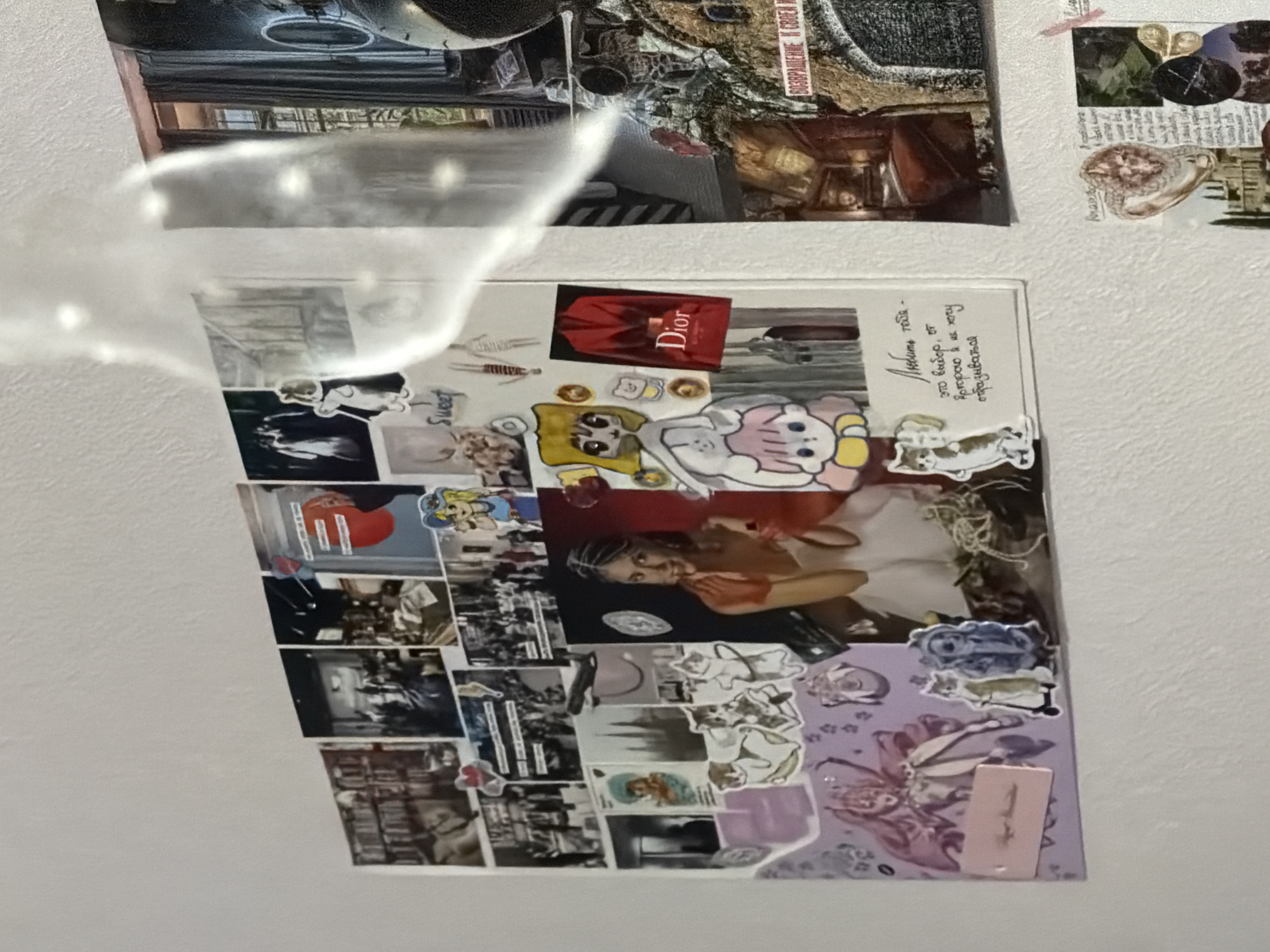 The image shows a collage of photographs and stickers on a white wall. The collage is arranged in a grid-like fashion, with some overlap between the elements. The photographs are of various subjects, including people, buildings, and abstract images. There are also several stickers, including cartoon characters, animals, and text. One of the stickers features a red Dior jacket with the Dior logo. Another sticker has a handwritten Russian text that reads "Люблю тебя - это выбор, от которого я не хочу отказываться" which translates to "I love you - it's a choice I don't want to give up."
To the right of the collage, there is another section of images and text, including a photograph of what appears to be an indoor scene with a bed and curtains, and a postcard with an inscription in Russian that seems to be a warning or a reminder, possibly related to "возвращение и своей игре," translating to "returning and your game." Below this, there is another small collage with various images including a heart-shaped object, a building with arches, and a circular image with a dark pattern.
The lighting in the scene suggests an indoor environment, possibly a room. There are no specific Moscow landmarks or clues visible in the image to pinpoint its exact location within the city. The overall impression is of a personalized decorative arrangement on a wall.