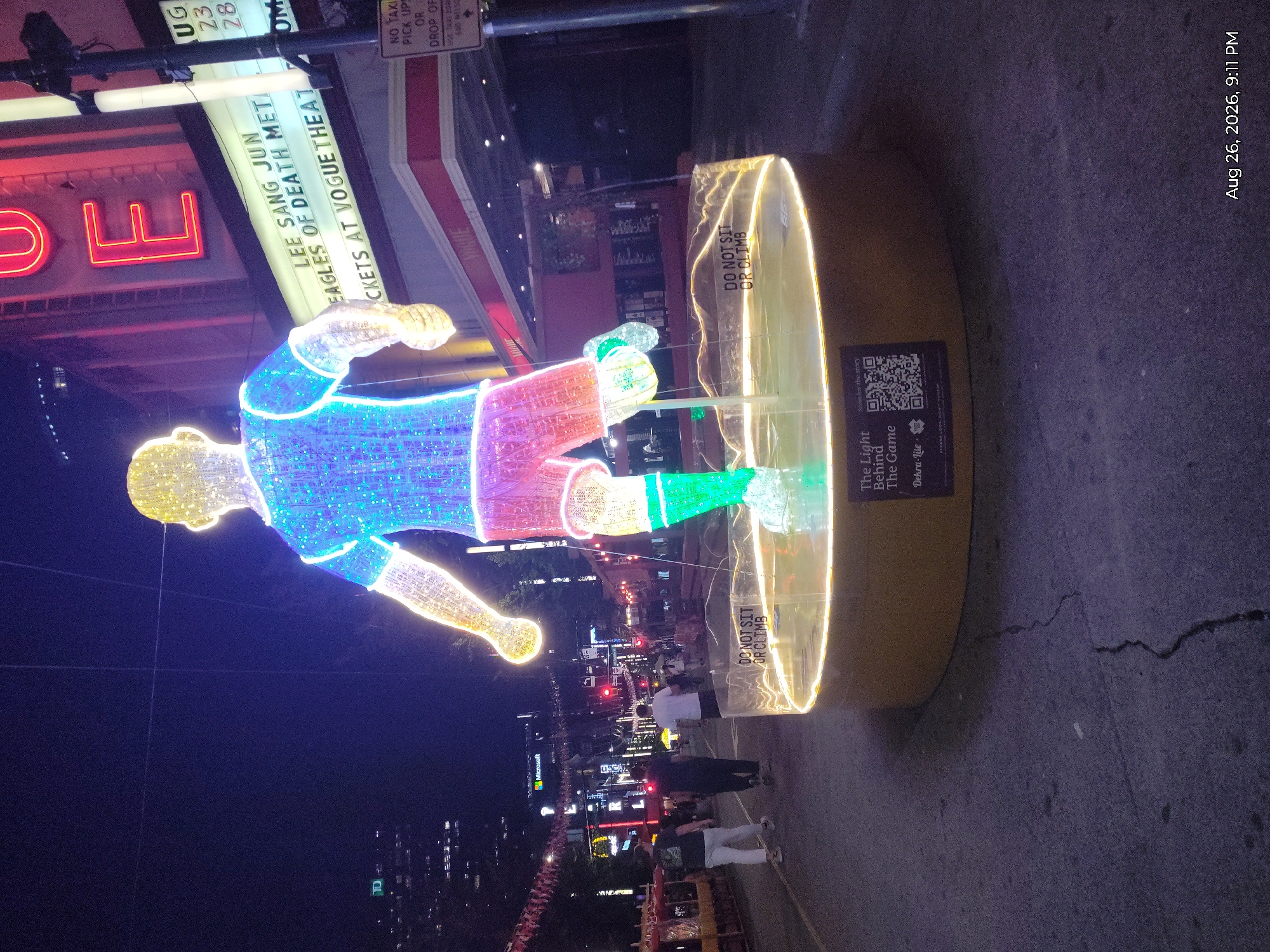 At night, a large illuminated sculpture of a soccer player stands in the middle of a city street, likely in Vancouver, Canada, given the presence of a "Vogue Theatre" sign and a "Microsoft" building visible in the background. The sculpture is made of colorful lights, with a yellow head and arms, a blue jersey, red shorts, and green socks. It stands on a circular, illuminated platform with a sign that reads "DO NOT SIT OR CLIMB." Several people are walking on the street in the background, and a movie theater marquee displays event information. The street is paved and appears to be a pedestrian area or a street closed for an event. The overall atmosphere is festive and urban.