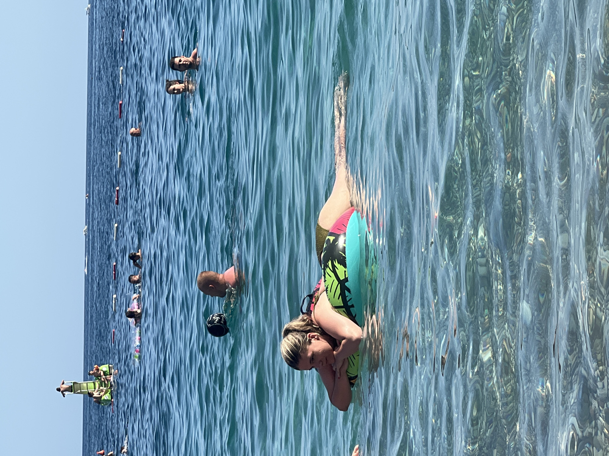 In a bright, sunlit scene at Kamenovo, Montenegro, several individuals are enjoying a day at the beach. The ocean, with its clear blue waters and gentle ripples, is the primary focus. The image captures a mix of people swimming, floating, and relaxing near the shore.
In the foreground, a woman is floating on an inflatable ring. The ring is decorated with a tropical palm tree pattern in vibrant green and black, with sections of pink and turquoise. The woman has blonde hair and is resting her head on her hands, looking towards the camera with a relaxed expression. Her body is partially submerged in the water, and her legs are extended, showing the colorful swimwear she is wearing.
To the left of the woman, a man is partially visible in the water, with his head and shoulders above the surface. He appears to be wearing a swim cap and has a focused expression. Further to the left, and in the background, more people are visible in the water. Some are swimming, while others are further out, appearing as small figures against the vast expanse of the sea.
Closer to the shore, a group of people are gathered around a bright green inflatable slide or raft. Some are sitting on it, while others are preparing to slide. This suggests a playful atmosphere and fun activities taking place.
The water's surface is a mosaic of light and shadow, with the sun's rays creating shimmering patterns and distorting the underwater view. The overall impression is one of a warm, pleasant day at the beach, with people enjoying the sea and the summer weather. There is no visible text in the image.