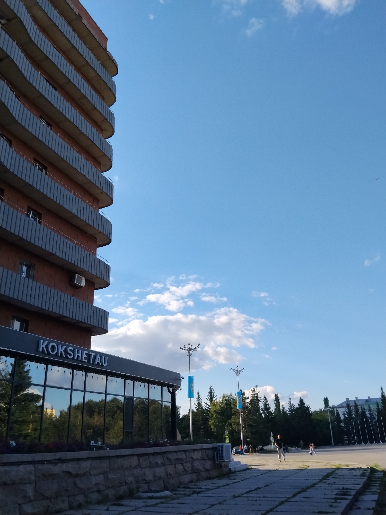 The image depicts a view of the city of Kokshetau, Kazakhstan, featuring a modern building with balconies and a glass-fronted lower level. The building's facade is primarily red brick with grey, curved balcony railings. The lower section of the building features large, reflective glass windows, showcasing a reflection of trees. Above the glass, the word "KOKSHETAU" is displayed in white lettering.
In the foreground, a paved area with a grid pattern of concrete slabs leads towards the building. A stone wall borders the paved area and the building's entrance. To the right of the building, a large open square is visible, dotted with trees and lampposts. Several individuals are present in the square. One person, a man wearing a dark t-shirt and light-colored pants, is walking across the square. Another individual, a woman in a light-colored outfit, is also present in the square. In the background, more trees and buildings are visible, and the sky is a clear blue with scattered white clouds. A bird is seen in flight in the upper right portion of the sky.