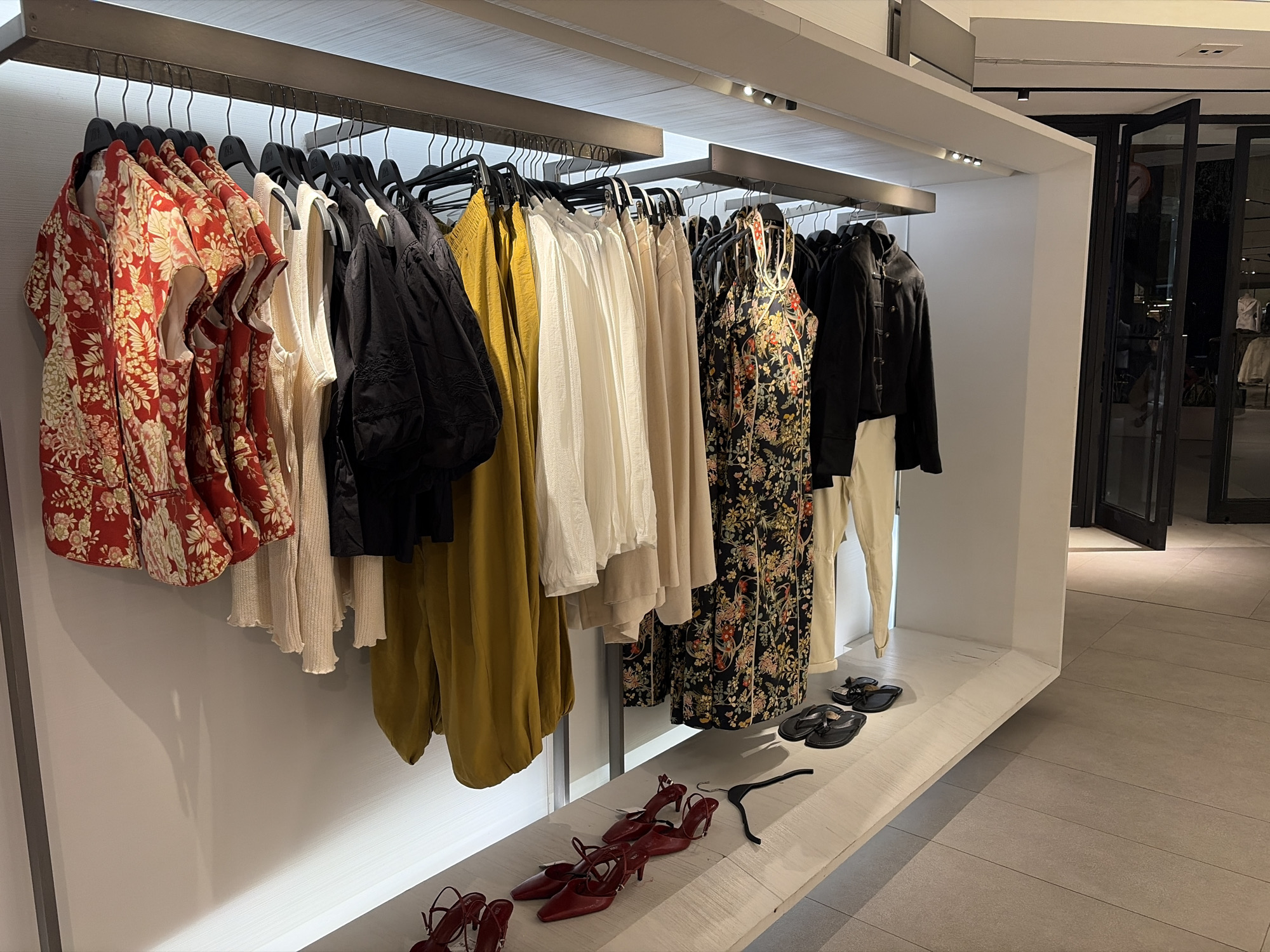 The image shows a clothing store display in Kuta, Indonesia. The scene features a rack of garments, including dresses and tops with floral patterns, solid colors like yellow, white, black, and cream, and a black jacket. On a shelf below, there are several pairs of red high-heeled shoes, two pairs of black flip-flops, and a black clothes hanger. The background shows a tiled floor and glass doors leading outside, indicating an indoor retail environment.