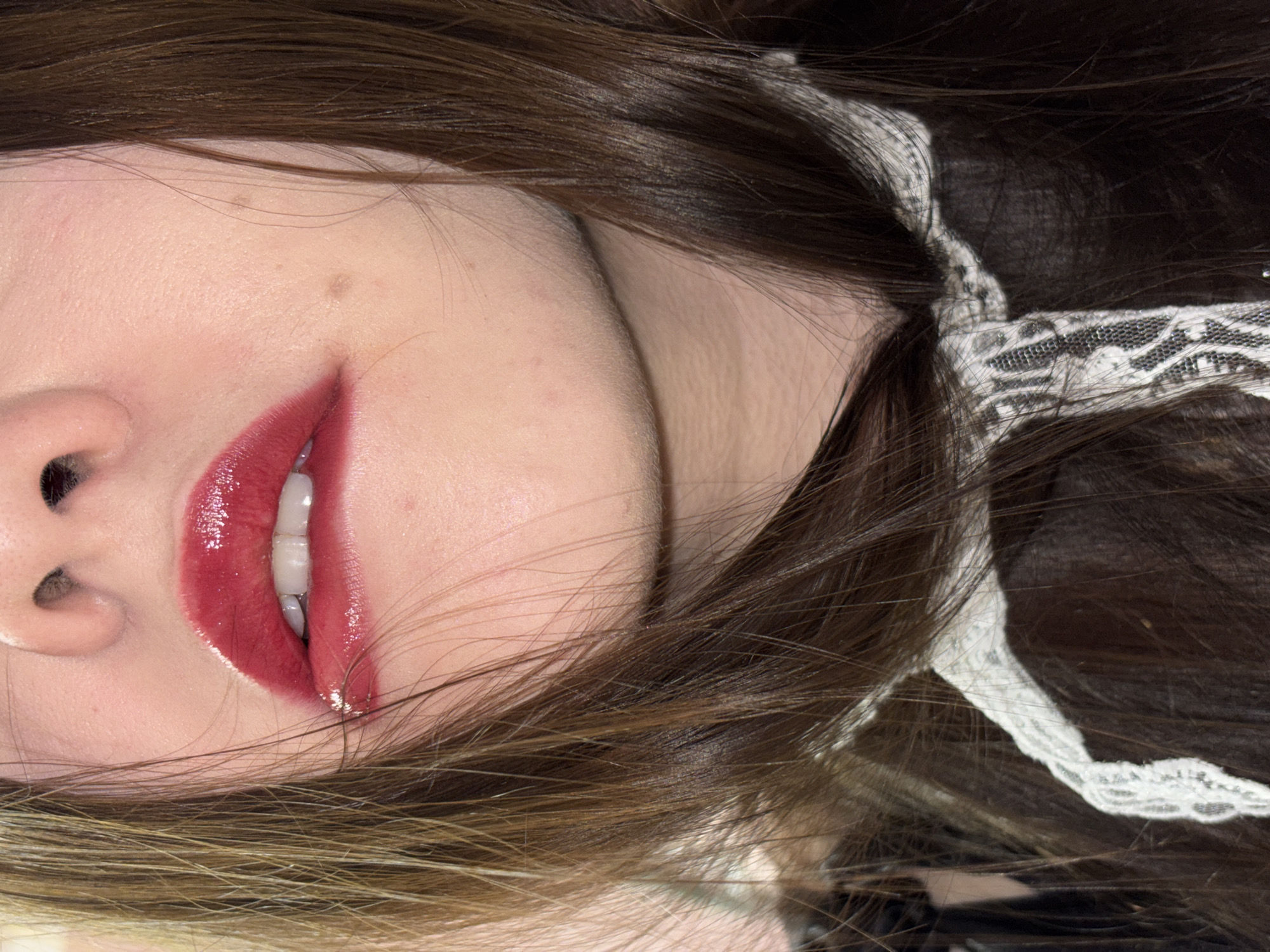 A close-up, angled portrait of a person's face, specifically from the nose and mouth up to the hairline. The individual has fair skin, with a visible mole on the cheek. The lips are coated in a glossy, deep red lipstick. Some teeth are visible. The person has brown hair, with blonde highlights towards the bottom. A white, lace-patterned hair tie is visible in the hair. There is no indication of the location being Perm, Russia.