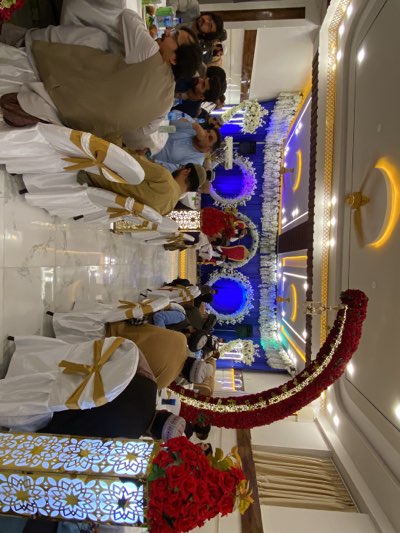 The image shows a wedding celebration taking place in an indoor venue. The room is decorated with white and gold chair covers, blue and white floral arrangements, and red floral garlands. Several men are seated in rows, wearing traditional clothing. Some of the men have headwear. The location is likely Kandahar, Afghanistan, based on cultural details and context.
