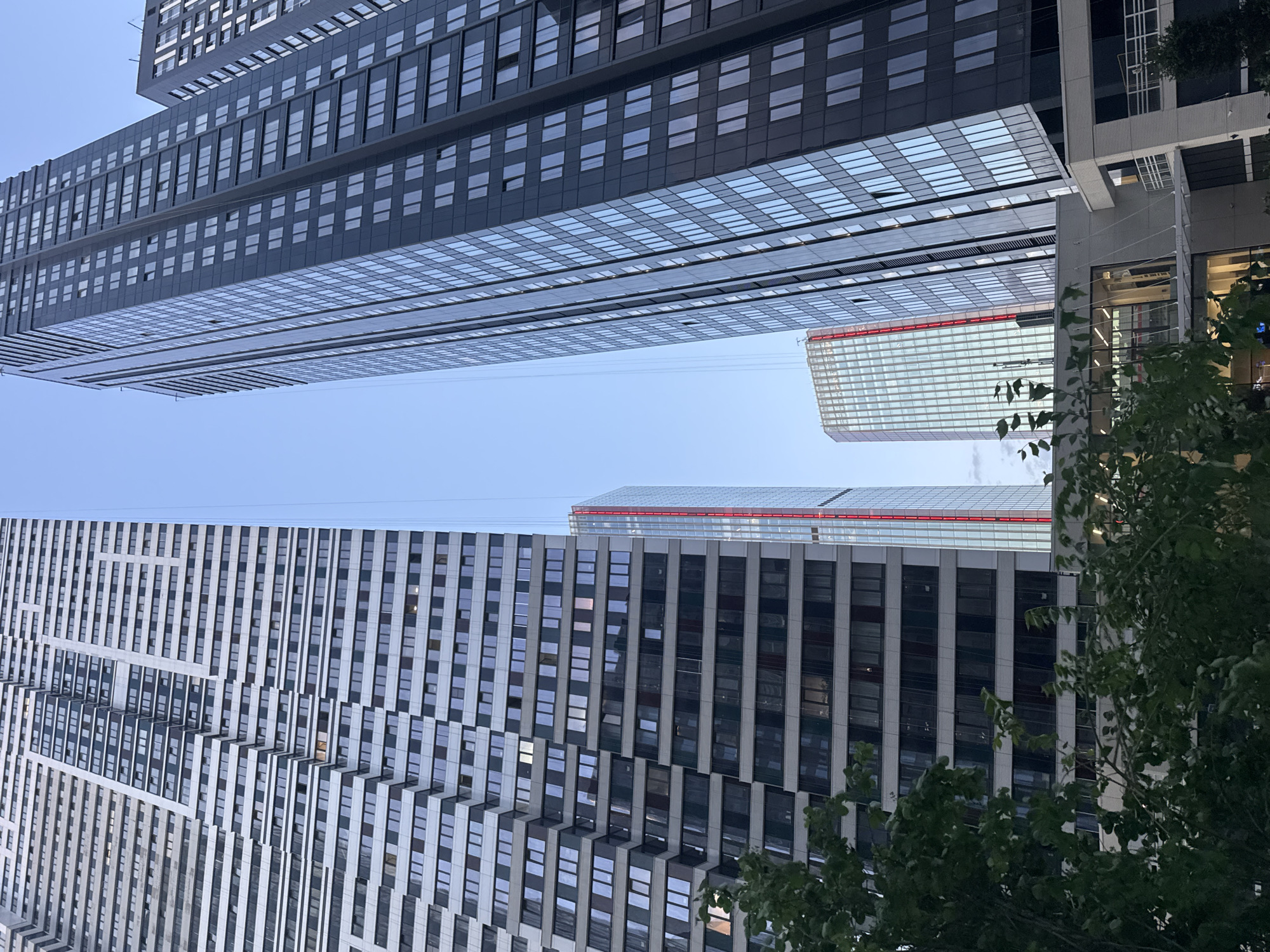 This media file presents an upward-looking view of several modern high-rise buildings against a clear, light blue sky, located in Khimki, Russia. The image is oriented such that the sky occupies the left side, and the buildings stretch horizontally across the frame.
Dominating the top section of the image is a large, dark-colored skyscraper. Its facade features numerous rectangular windows arranged in uniform grids. The underside of this building, facing the sky, is composed of reflective glass panels that mirror the blue sky. A prominent cantilevered section extends from this structure.
Across the bottom section of the image lies another substantial, modern building. This one has a lighter, possibly white or light grey facade, adorned with a distinctive, complex pattern of alternating vertical and horizontal elements integrated with windows, creating a textured appearance.
In the middle distance, positioned between these two primary structures, a third contemporary building is partially visible. It exhibits a light-colored, likely glass and white exterior, and is topped with a notable horizontal red stripe or lighting element.
The background is a uniformly clear, light blue sky, indicating fair weather. In the bottom-right corner, green leaves and branches from trees or bushes partially frame the architectural scene. There are no visible people or discernible activities taking place, and no text is readable on any of the structures. The overall impression is one of modern urban architecture viewed from an unconventional angle.