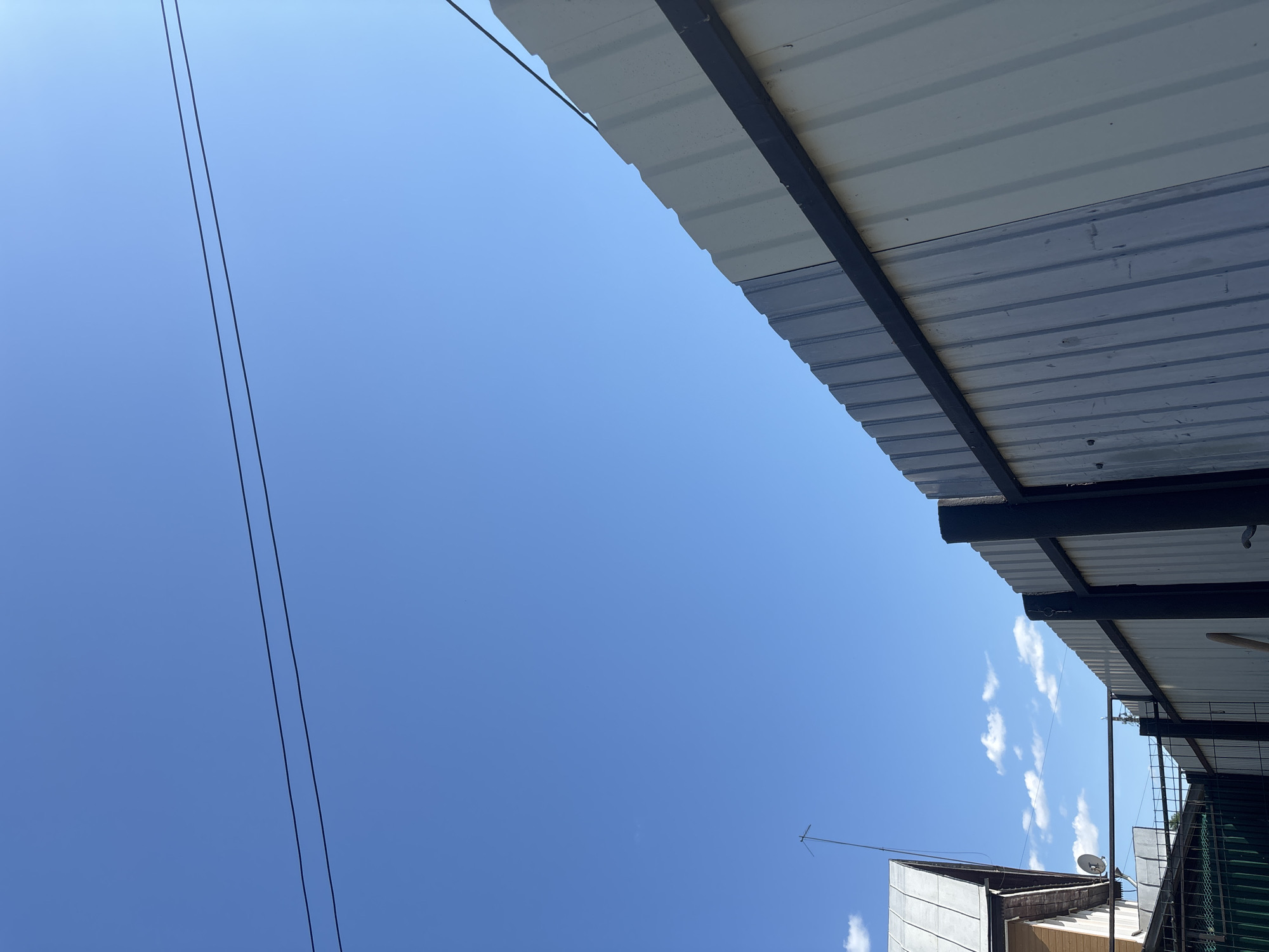 The image depicts a bright, clear blue sky dominating the upper portion of the frame, with two slender, parallel utility lines stretching horizontally across the upper-middle section. Scattered white cumulus clouds are visible in the lower-left background. A tall, light-colored corrugated metal fence, supported by dark vertical posts and a horizontal crossbeam, diagonally bisects the lower right part of the image, angling upwards from left to right. The fence shows signs of minor wear and some variation in panel color, ranging from off-white to light grey. In the lower-left background, a portion of a building, likely a single-story house, is visible, featuring a gabled roof made of a grey, possibly metallic, material. A tall, slender antenna with cross-elements extends vertically from the house's roof, and a small satellite dish is affixed to its side. Below the house, segments of a darker, possibly green, mesh fence are partially visible. The scene is bathed in bright daylight, indicative of a sunny day in a suburban or rural area of Semyonov, Russia. No individuals are present in the frame.
