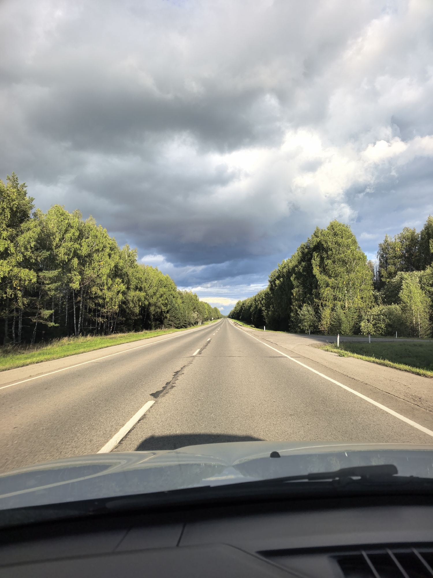 The image depicts a frontal view from inside a vehicle driving on a paved road through a wooded area. The road stretches into the distance, flanked by dense green trees on both sides. The sky is filled with dramatic, dark gray clouds, suggesting impending rain or a recent storm. The sun is partially visible, casting light on the clouds and illuminating parts of the landscape. The foreground shows the car's dashboard and windshield wipers. There are no people or animals visible in the scene. The location is identified as Shabury, Russia.