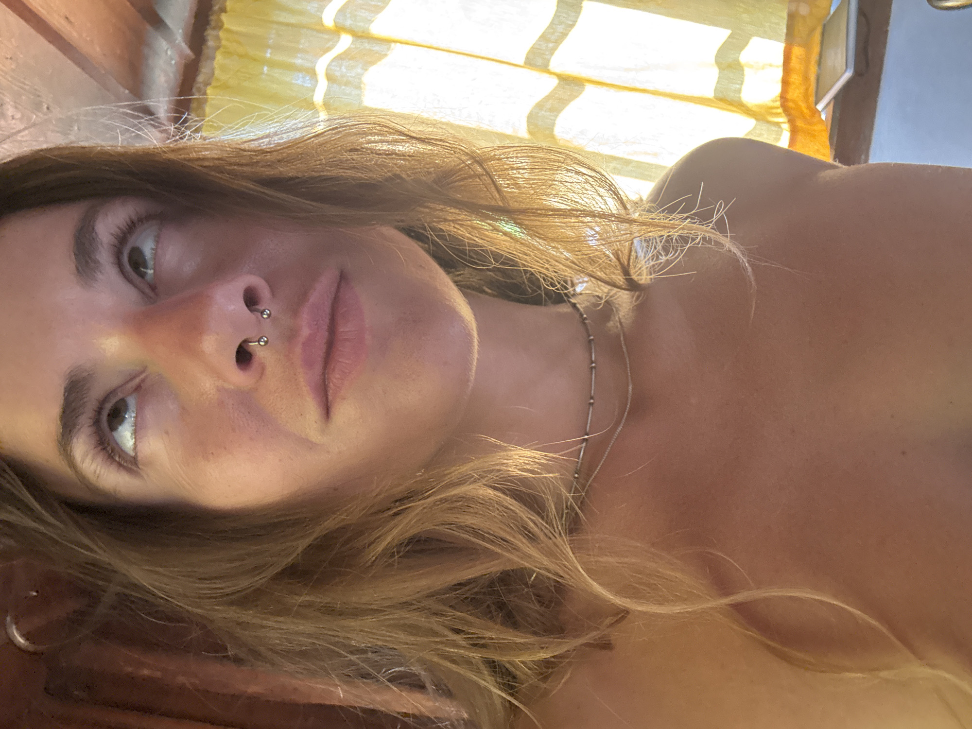 This is a close-up, upside-down, indoor selfie of a person with light brown hair. The person is looking towards the camera with their head tilted. They have a septum piercing and a delicate silver necklace. The background is slightly blurred, showing what appears to be yellow blinds or curtains illuminated by bright sunlight, suggesting it is daytime. The lighting creates a warm glow on the person's skin. The scene appears to be a relaxed, personal setting. There is no visible text in the image.