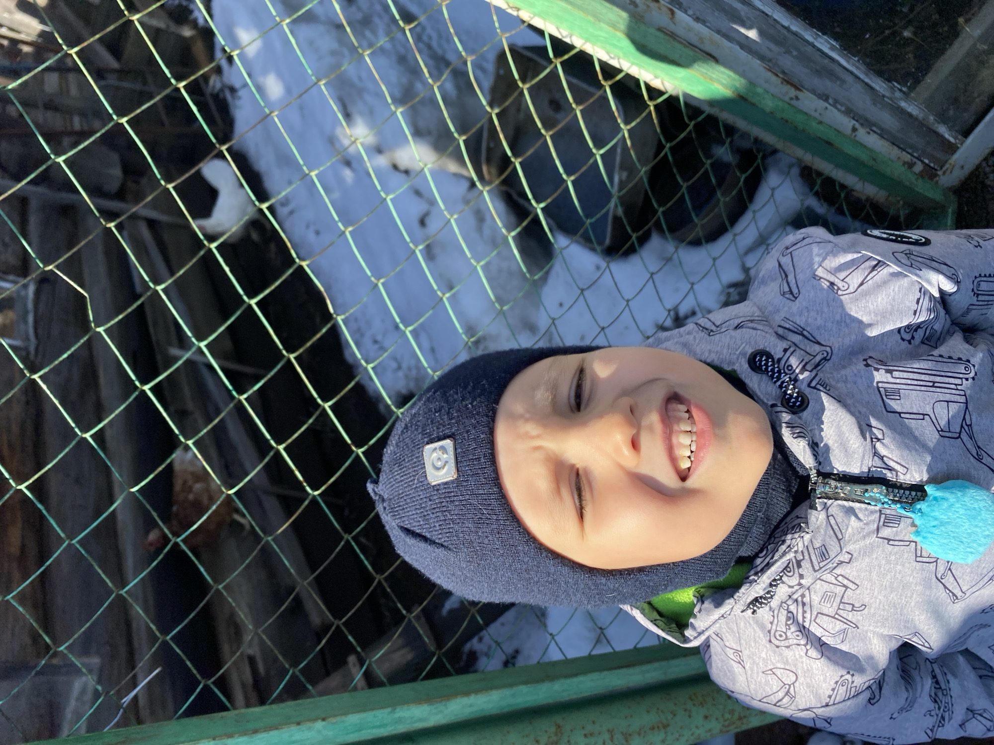 A young boy wearing a dark blue knit hat and a light gray jacket patterned with drawings is positioned near a wire mesh fence with green framing. The background shows patches of snow and dark, possibly wooden, structures visible through the fence. The boy is smiling and appears to be facing the camera. The photograph appears to have been taken in daylight, possibly outdoors. There is no information within the image or prompt connecting it to the city of Noviy, Russia.