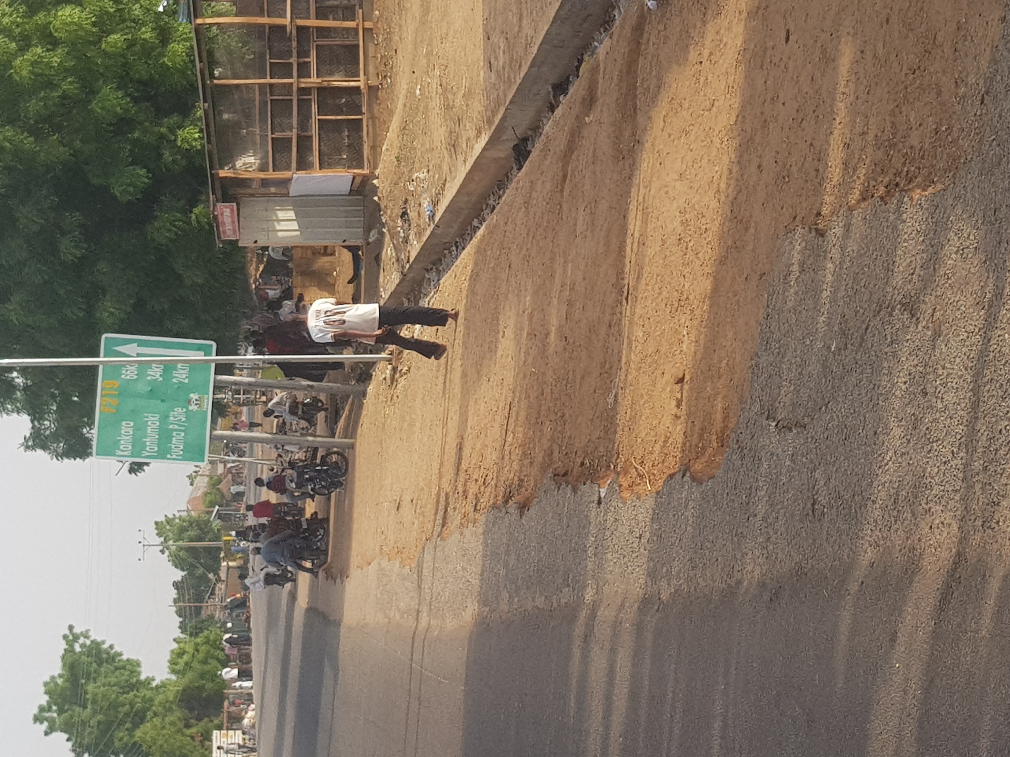 The image depicts a scene in Dabawa, Nigeria, featuring a road alongside a sandy shoulder. A road sign indicates directions to Kankara (66 km), Yanlumoki (34 km), and Fudmo P/Site (24 km). Several people are present, some of whom are riding or standing near motorcycles. A small, wooden structure or shop stands near the road. The environment consists of trees, telephone poles, and buildings in the background, creating a busy roadside setting.