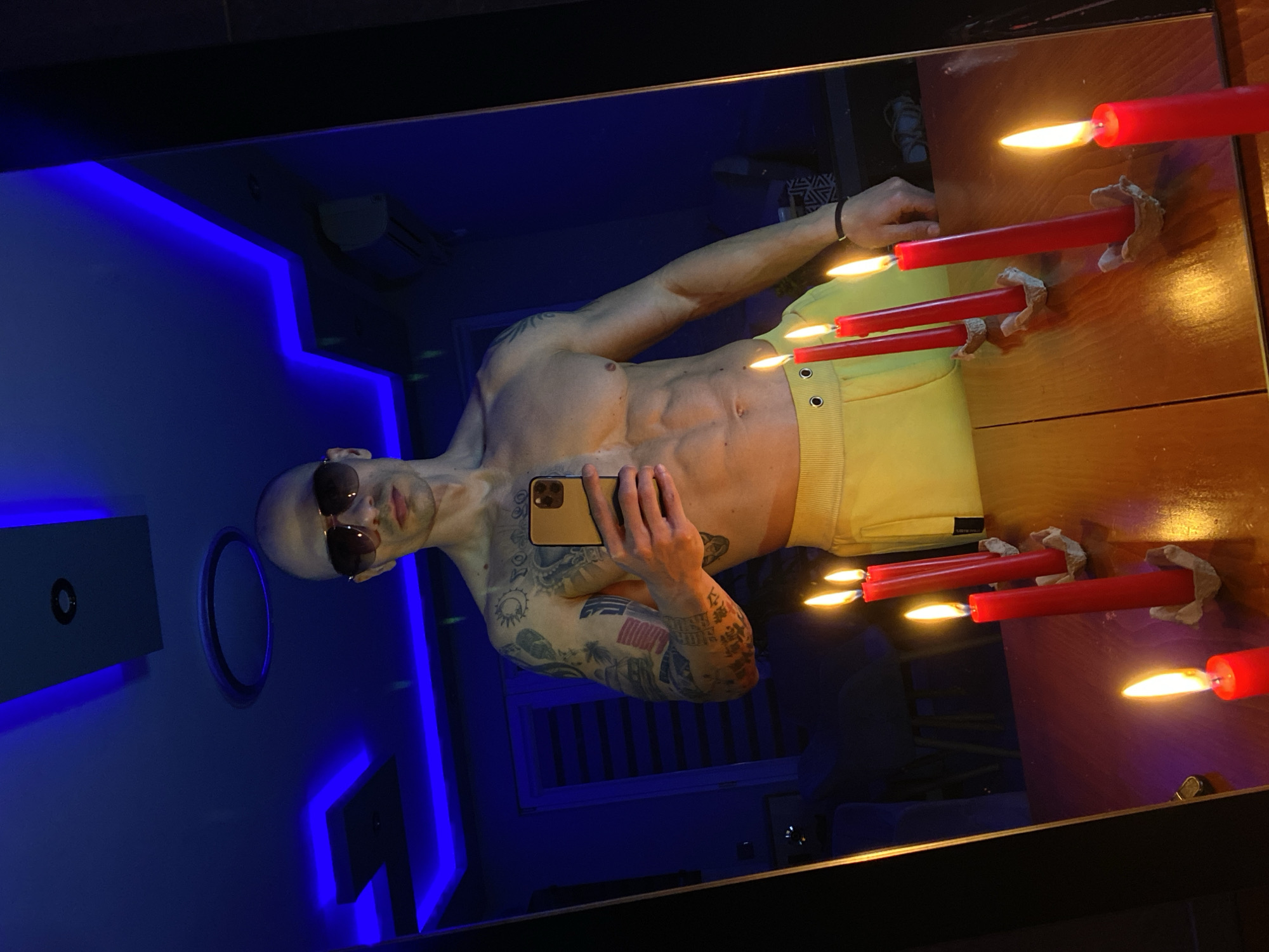 A shirtless man with a shaved head and sunglasses is taking a selfie in a mirror. The man has tattoos on his left arm. He is wearing yellow shorts. The background is a room with blue neon lighting. There are red candles on the surface in front of the mirror. The room contains a small round wall-mounted object and a wall-mounted black square object. It is not possible to determine the location in Pleven, Bulgaria from the image.
