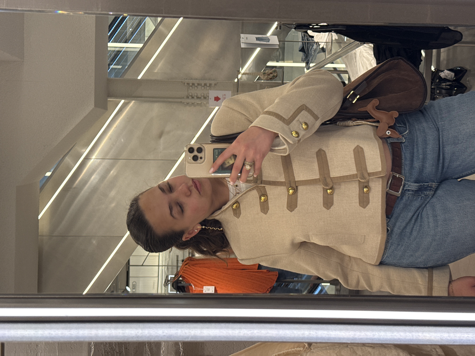 A young woman is taking a selfie in a store mirror. The mirror reflects the store's interior, revealing clothing racks with various garments, including an orange sweater. The woman is wearing jeans, a beige military-style jacket with gold buttons, and a brown shoulder bag with a whimsical bird-shaped detail. She holds her iPhone with a decorative case, and she's wearing earrings and rings. The mirror's reflection also captures part of the store's ceiling with architectural lighting. The exact location within Milan is not discernible, but the environment is that of a retail clothing store.