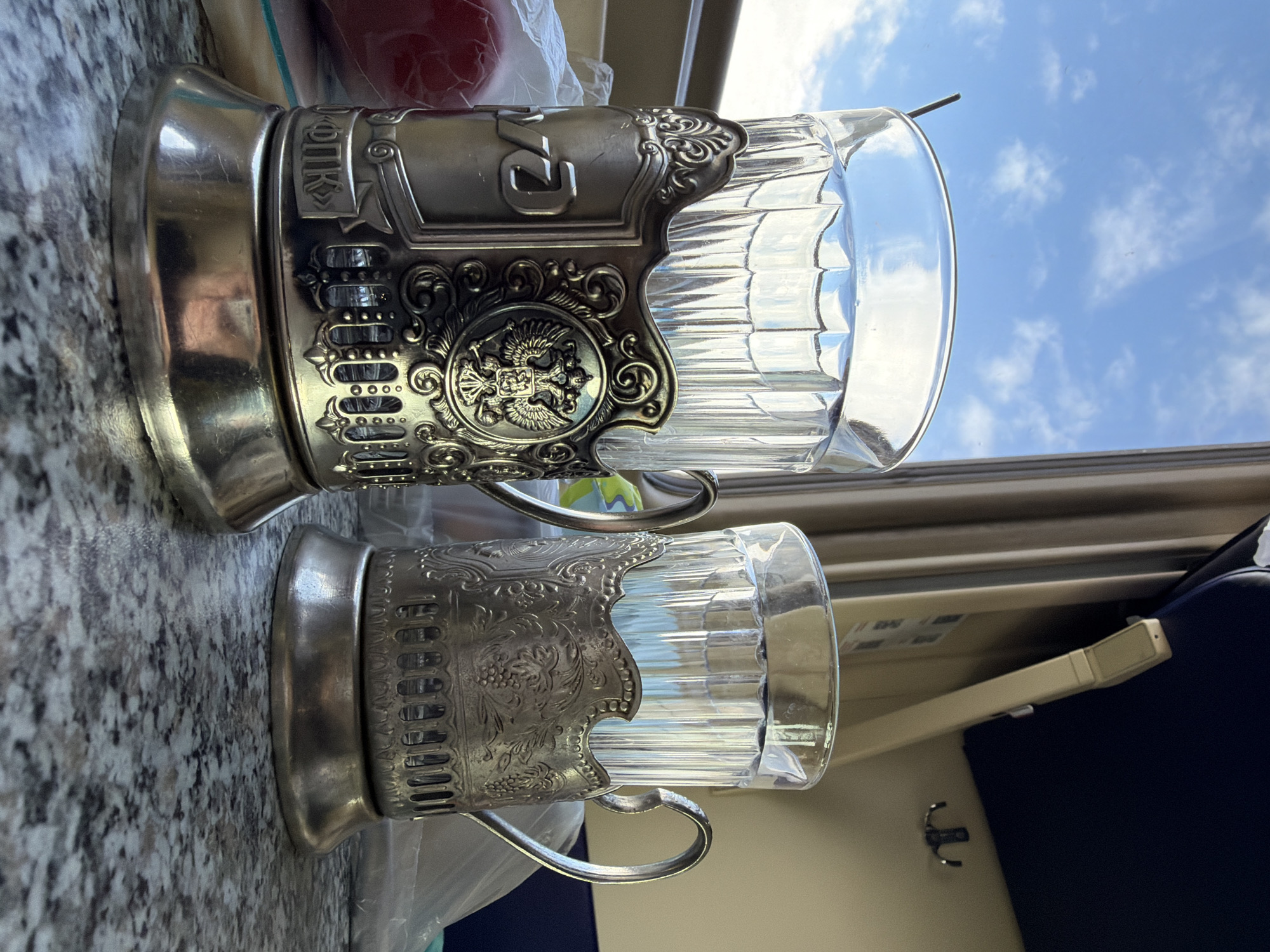 Two ornate silver-plated glass holders, typical of Russian train travel, rest on a surface. The top holder is more elaborate, featuring the Russian double-headed eagle emblem. A portion of a window showing a blue sky with clouds is visible behind the holders. In the background, train interior elements are visible, like a panel, a folding tray, and a blue seat. It's likely these glass holders are inside a train in or around the city of Berezovyi, Russia.