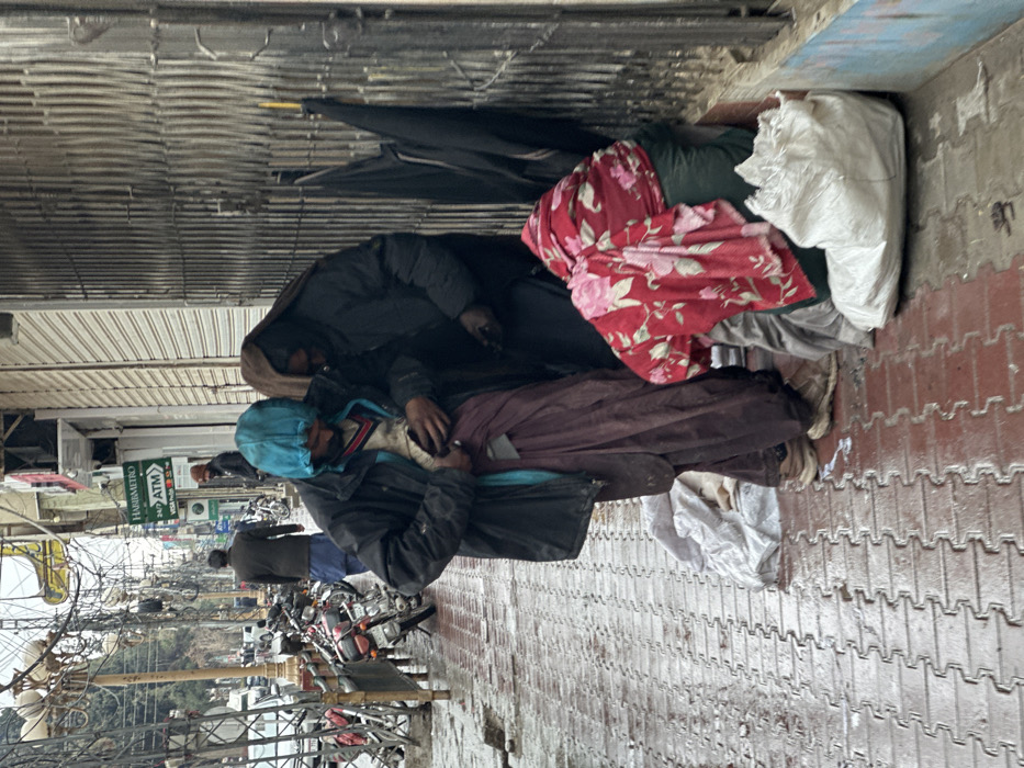 This image depicts a wet urban street scene in Quetta City Tehsil, Pakistan. The red-brick sidewalk and asphalt road are visibly damp, indicating recent precipitation.
In the foreground, three individuals are present. On the left, a male subject stands, wearing a dark hooded jacket with a blue hood, a striped sweater, and maroon/brown baggy trousers (shalwar) over light-colored sneakers. He is holding a white mobile phone at waist level, appearing to operate it with both hands. To his immediate right and slightly behind him stands a second individual, largely obscured, dressed in dark clothing, including a head covering. They are oriented towards the first individual. Further right, a third individual is seated on the ground, leaning against a metal storefront security shutter. This person is largely covered by a red blanket with a white floral pattern. A white plastic bag rests on the ground in front of them, and three black, folded umbrellas lean against the shutter to their right. A white cloth item is on the ground near the first individual's feet.
The mid-ground features a line of parked motorcycles on the wet sidewalk, followed by a street with additional pedestrians and distant vehicles. Buildings line the street, including one with a green "HABIB METRO ATM" sign visible. Overhead, a dense network of black electrical or communication wires crisscrosses the sky.