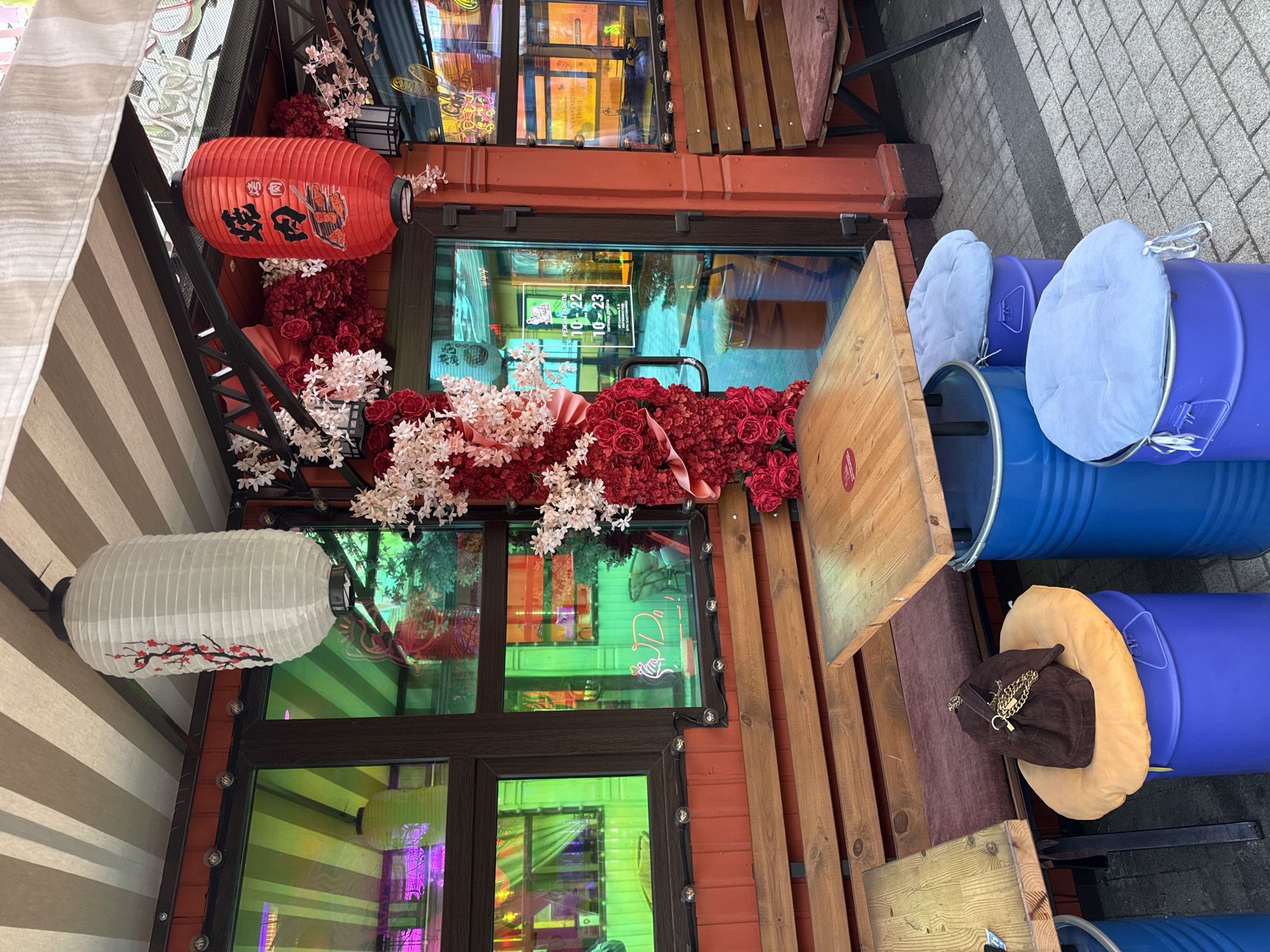 The image depicts the exterior of a restaurant in Krasnodar, Russia. The restaurant has a vibrant red facade and is decorated with Japanese-style lanterns and artificial red and pink flowers. The windows are tinted with a green and purple iridescent film, creating a colorful reflection of the surroundings. There are outdoor seating areas with wooden tables and benches, and unusual seating options made from blue barrels topped with cushions. A brown handbag with a gold chain is placed on one of the barrel seats. The ground is paved with grey cobblestones. The overall impression is a lively and eclectic establishment.