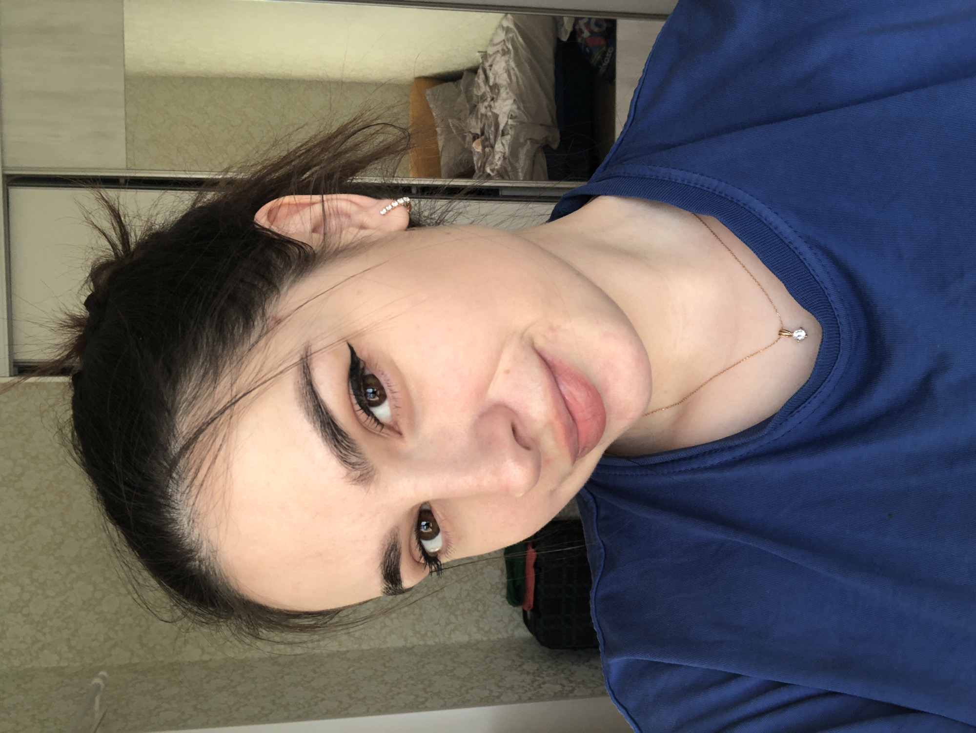 A woman with dark hair pulled back and dark eyeliner wears a blue t-shirt and a diamond necklace. She is wearing a curved earring with multiple small diamonds. The background includes a mirror, and a blurred interior room with objects that are difficult to discern. The location is unknown and there is no information suggesting the city of Noviy, Russia.