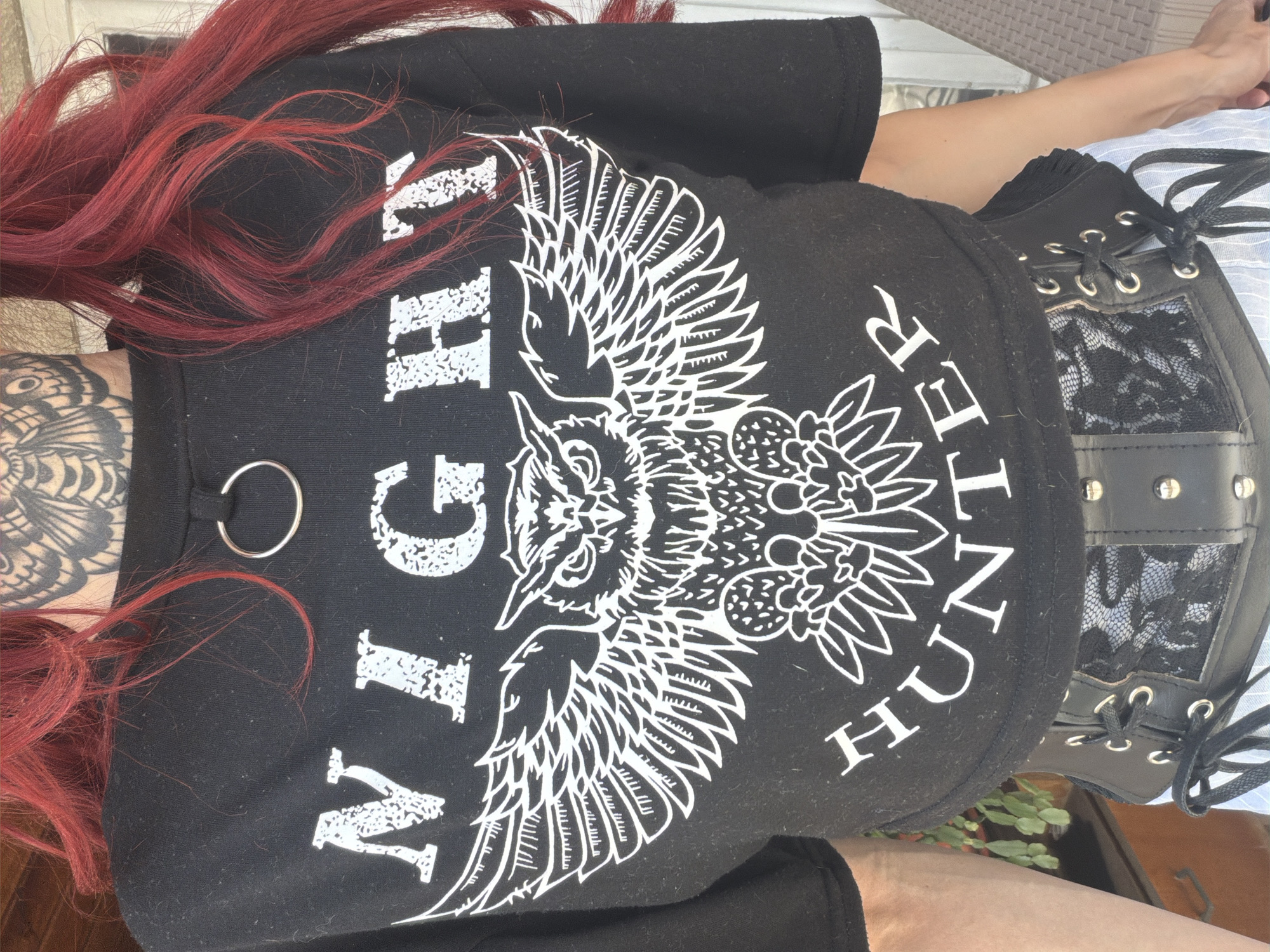 An individual with bright red hair and facial tattoos stands outdoors, cropped from the chest up. They are wearing a black t-shirt with a white graphic of an owl and the words "NIGHT HUNTER" printed on it. The t-shirt has a silver ring detail at the neckline. Below the t-shirt, a black corset with lacing and silver grommets is visible, worn over a white and blue striped garment. The background is blurred but appears to be an outdoor setting, possibly a patio or garden. The image does not provide enough information to determine the location in the city of Svilengrad, Bulgaria.