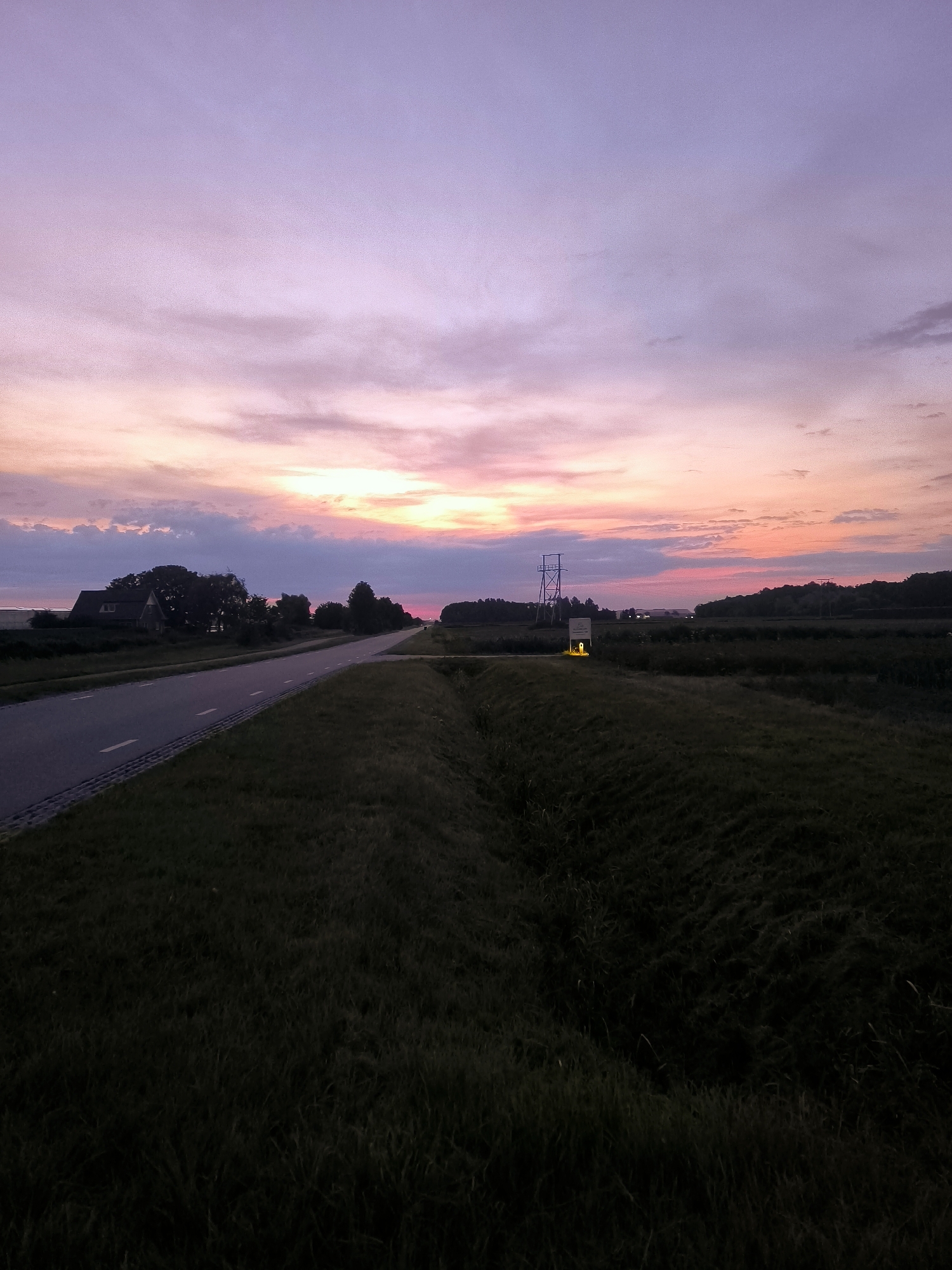 This is a wide-angle, eye-level shot of a paved road curving to the left, bordered by a grassy verge and a field of grass. The road is empty and has white dashed lines indicating lanes. In the distance, on the left side of the road, is a house with a dark roof and a few trees. On the right side of the road, further in the distance, is a tall, metal structure with a yellow light at its base. The sky is filled with clouds that are illuminated by a sunset, displaying shades of purple, pink, and orange. The overall environment appears rural, and the scene depicts a tranquil evening in Andijk, Netherlands.