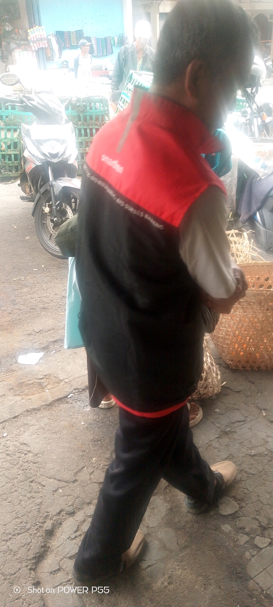 In an outdoor urban setting, a person walks with their back to the camera, wearing a black vest over a white shirt and dark pants. The vest has a red collar and a red stripe along the bottom hem. Text is visible on the vest, reading "smadhen" and "NO SUGES". A motorcycle is parked to the left of the frame. In the background, a building with a light blue facade is visible, with various goods displayed on shelves. Other people are present in the background, including one wearing a cap and another with a white cap. The ground is paved with asphalt and cracked in places. Two woven baskets are on the ground to the right of the walking person. The image is watermarked "Shot on POWER P55".