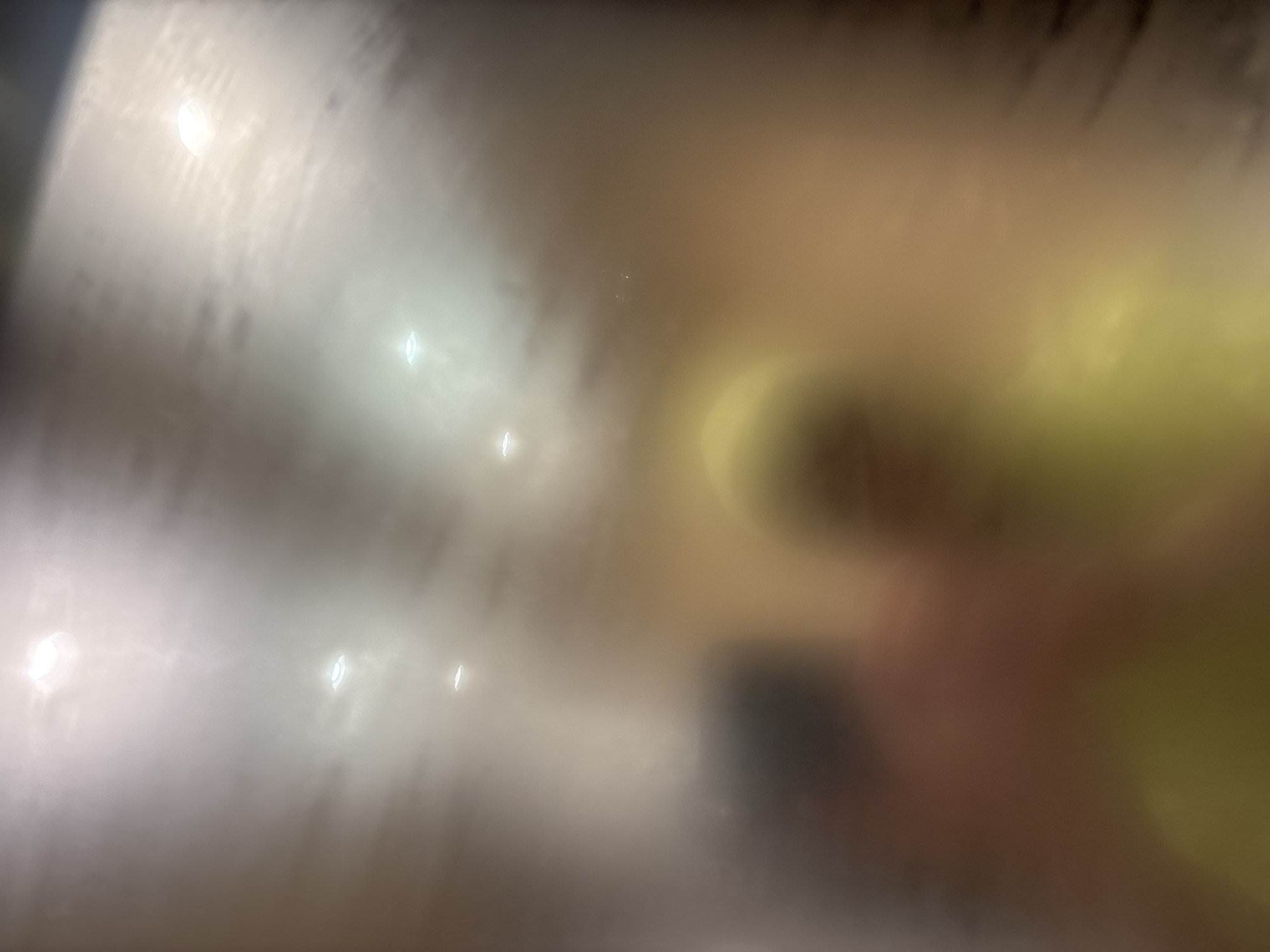The image is blurry, but it appears to depict the interior of a building. The left side of the image has light reflections that may be from lights on the ceiling. There are indistinct shapes that may indicate the presence of people and objects. The location is unknown.