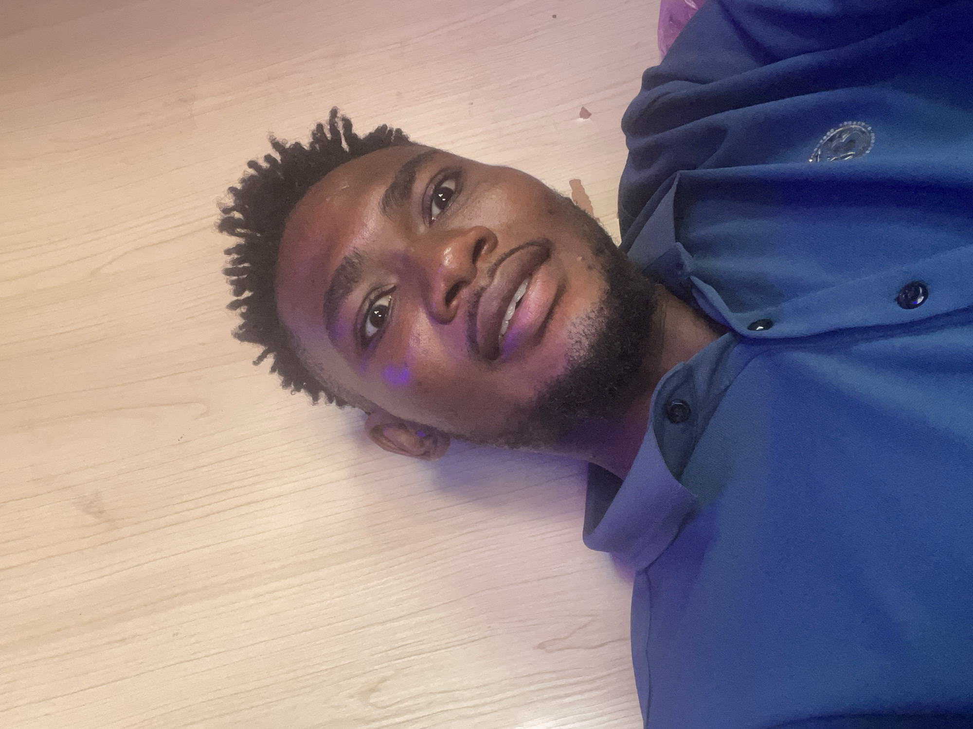 A young man is lying on a light-colored, horizontally-aligned, wooden floor in Samaru, Nigeria. He is wearing a dark blue collared shirt with buttons. The man has short, dark, coiled hair, a dark beard, and brown skin. There appears to be a purple light source casting a hue on the left side of his face.
