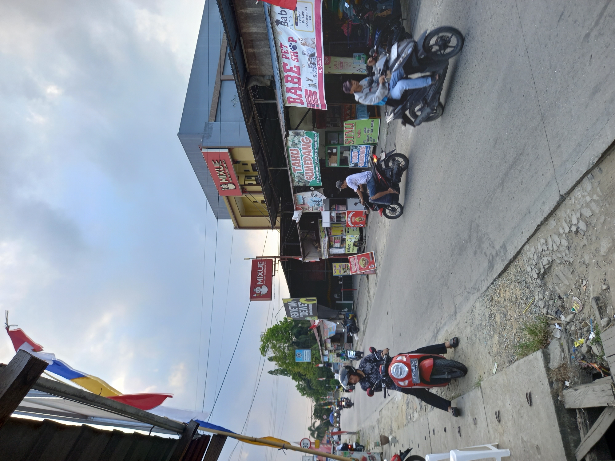 The image captures a street scene in Samarinda, Indonesia, with a focus on a road lined with businesses and pedestrian activity. In the foreground, a person is seated on a red scooter parked on the side of the road. The scooter has a license plate that reads "KT 5768 SZ". Further down the road, two men are riding motorcycles, with the rider on the left on a red motorcycle and the rider on the right on a black motorcycle, both moving away from the viewer.
The buildings lining the street exhibit a mix of commercial signage. Notably, there are prominent red signs with "MIXUE" branding, indicating ice cream and tea shops. Other signs advertise services like "TAHU SUMEDANG" (a type of fried tofu) and "BABE PET SHOP". The architectural style appears to be functional and utilitarian, with awnings and simple building facades. The street itself is paved, with some signs of wear and tear, including debris and cracks.
The environment is characterized by a cloudy sky, suggesting a potentially overcast day. Several utility poles with wires are visible, typical of urban infrastructure. Trees are present in the background, adding a touch of greenery to the scene. The overall impression is that of a bustling, everyday street in a commercial area of Samarinda.