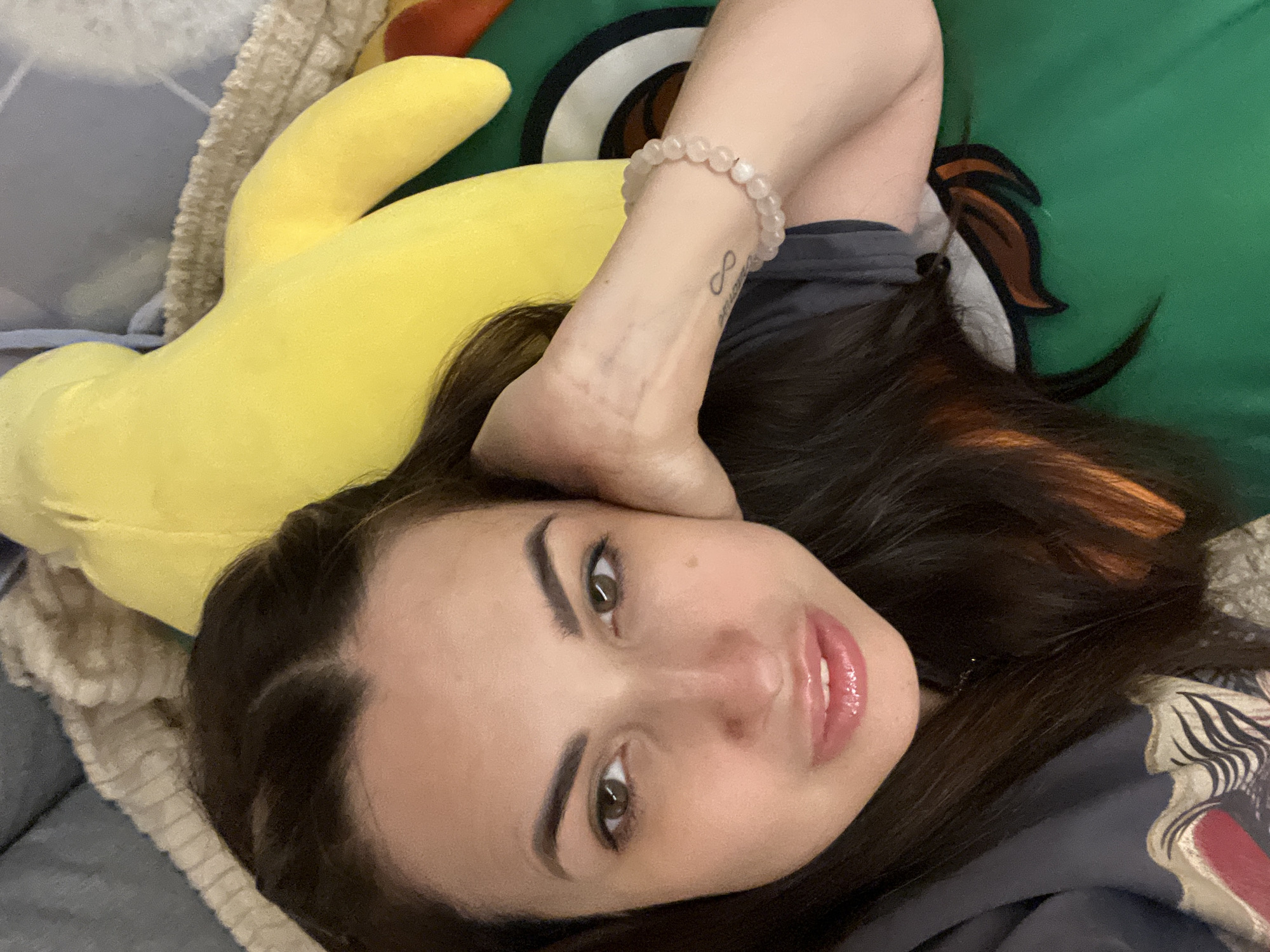 An overhead, close-up shot shows a woman with dark hair and brown eyes resting her head on her hand. She is wearing a grey t-shirt and a bracelet made of pale pink beads. Next to her is a large, yellow stuffed animal, possibly a banana, and a green object with cartoonish eyes. The background consists of a gray blanket and a beige textured fabric.