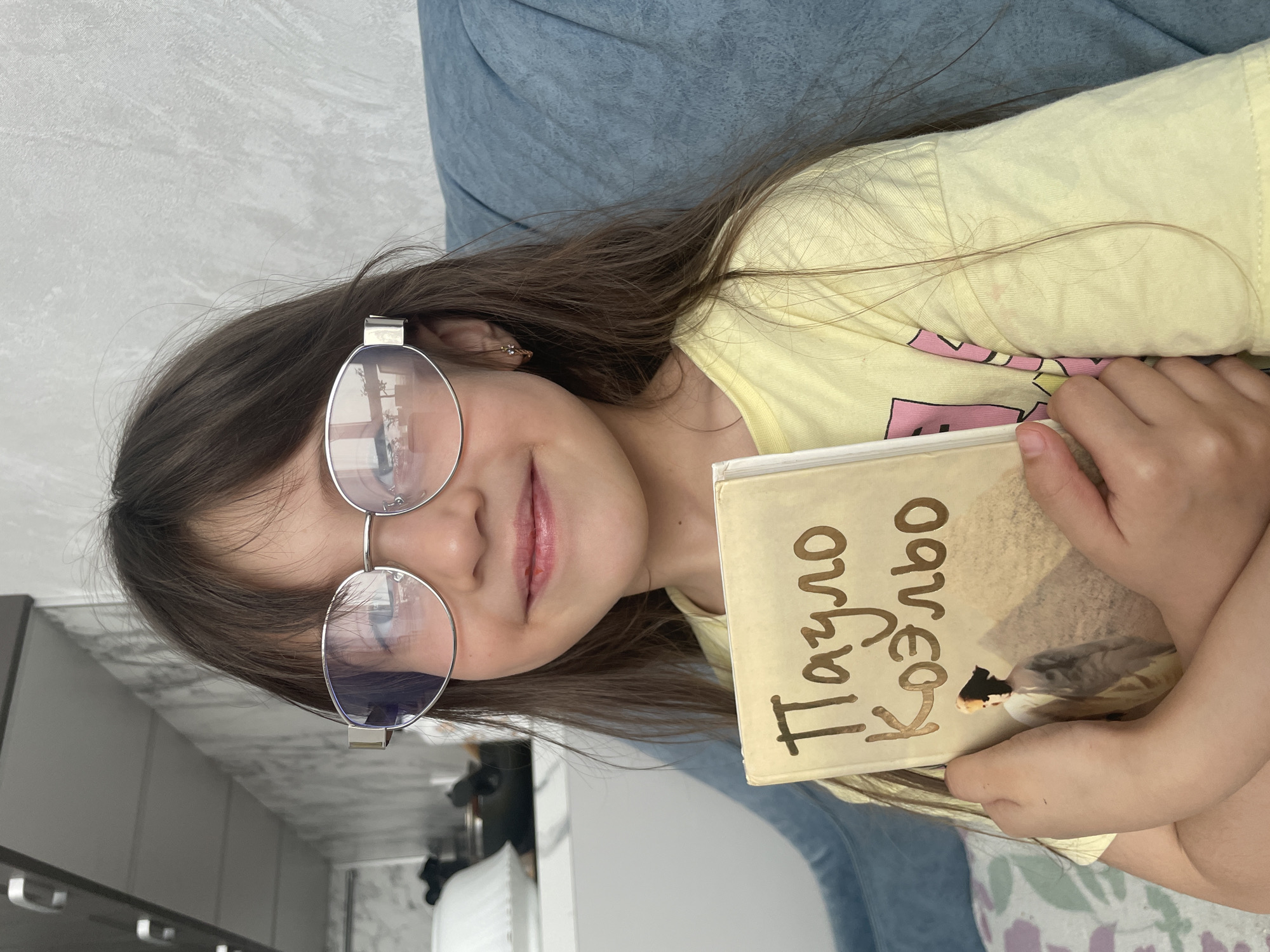 A young girl with long brown hair and silver-rimmed glasses is smiling while holding a book. The book's cover is tan and has the name "Paulo Coelho" written in Cyrillic script. She is wearing a yellow t-shirt and is partially reclined against a blue surface, possibly a couch or cushion. In the background, there is a glimpse of a room with light walls and a dark cabinet. It is not possible to determine if this image was taken in Izhevsk, Russia.