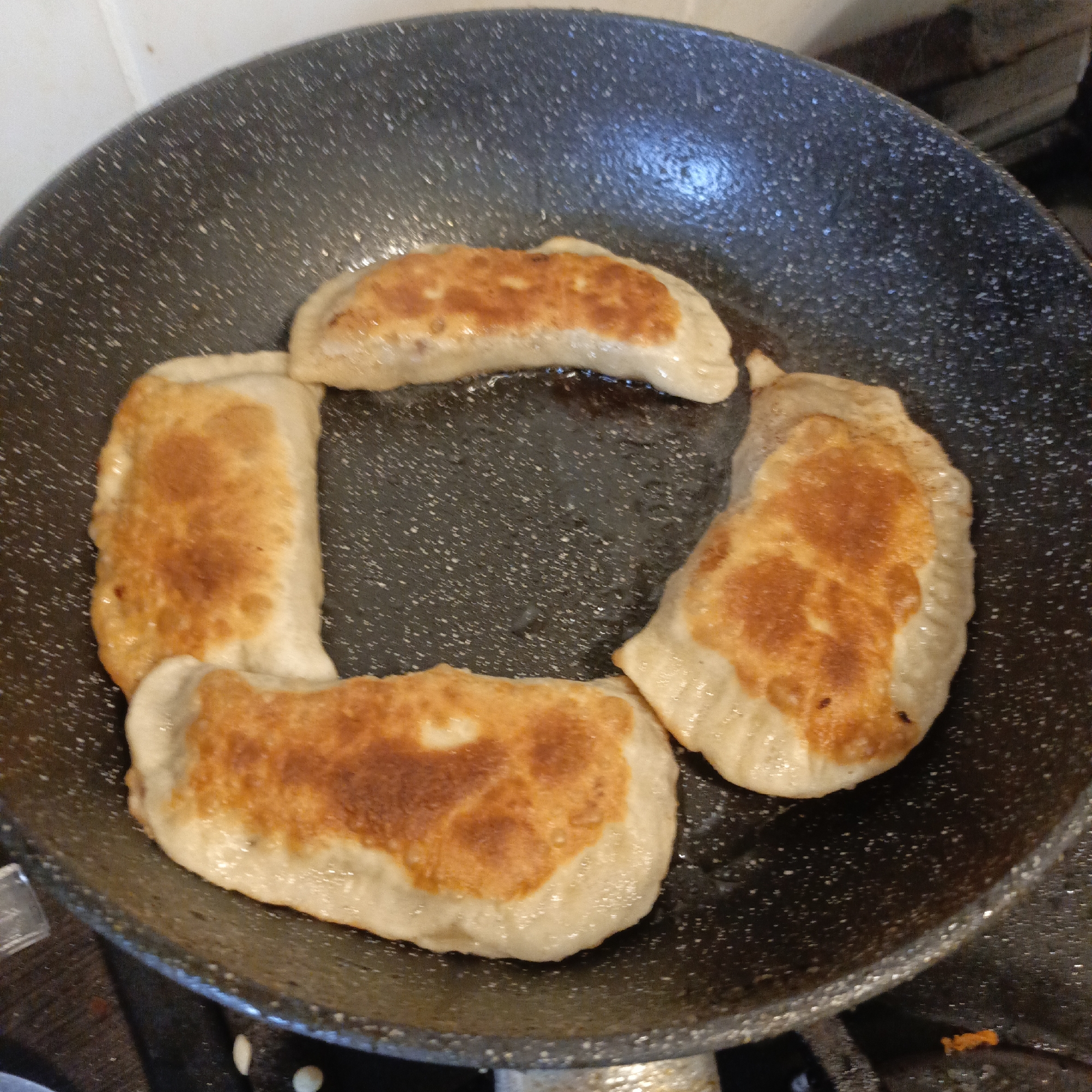The image shows a black frying pan containing four fried pierogi, arranged in a rough circle. They are golden brown on one side, suggesting they have been cooking in oil. The pan is on a stovetop, which can be seen in the bottom part of the image. The location of the image is Unknown, Unknown.