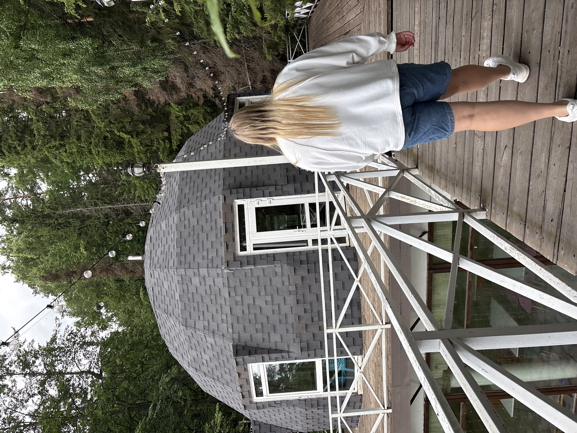 A blonde-haired person wearing a white sweatshirt and denim shorts walks away from the camera on a wooden bridge. The bridge has white railings and leads towards a grey geodesic dome-shaped building with several windows and a door. The building is surrounded by lush green trees. String lights are visible overhead. The scene appears to be in an outdoor, natural setting, possibly a resort or park.