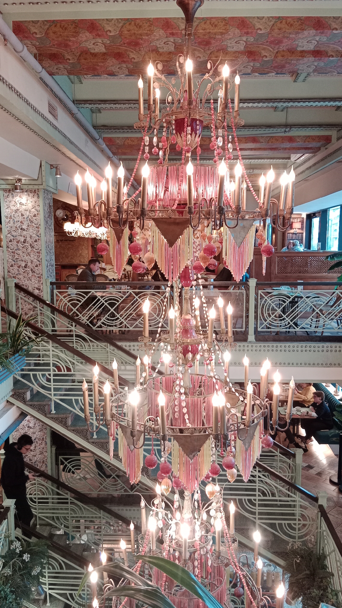 Captured from an elevated angle, the interior of a building, most likely in the city of Unknown, Unknown, is revealed. A grand, multi-tiered chandelier with numerous faux candle lights dominates the central composition. The staircase features a distinct design, and people are dispersed throughout the interior: a person is going down the stairs, some are seated at tables, and a few are positioned behind a railing on an upper level. The ceilings are ornamented with patterns.