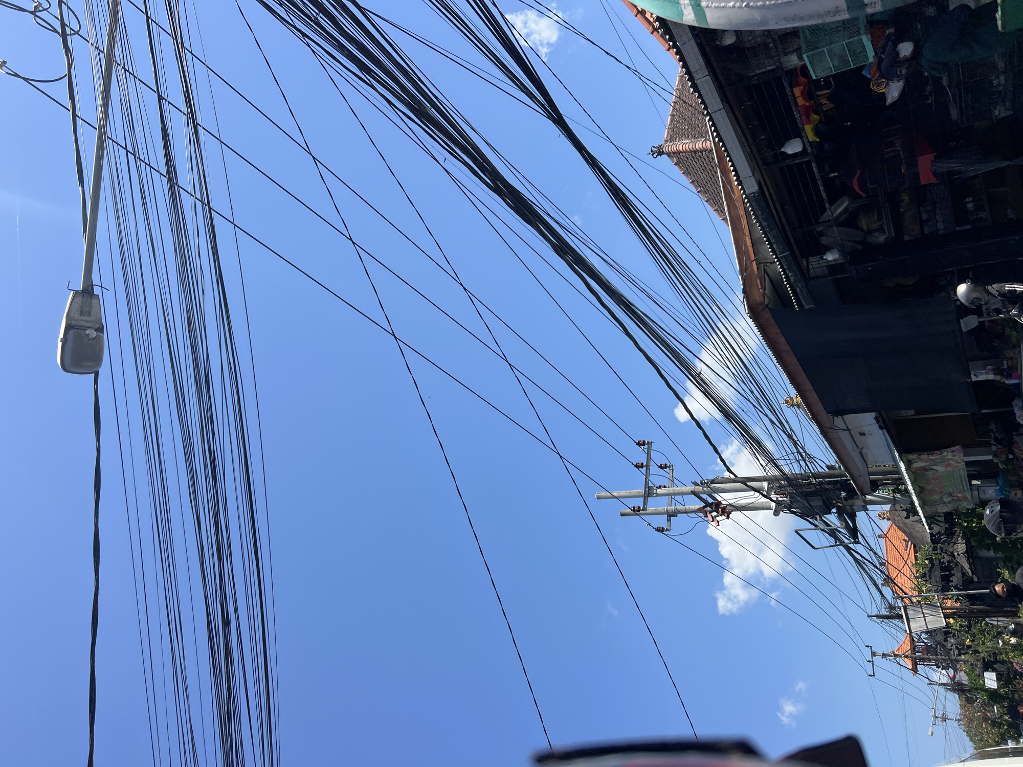 A low-angle, full shot captures a bright, clear blue sky filled with a dense network of black electrical wires crisscrossing the frame. A street lamp is visible on the upper right, extending from a pole. Below the wires, the rooftops of buildings are visible, with terracotta tiles on some and corrugated metal on others. One building appears to be a shop with an open front, displaying various items on shelves, including what appear to be birdcages. Several utility poles with transformers and insulators are also present. In the foreground, parts of vehicles, possibly motorcycles, and a person wearing a helmet are visible. The scene is likely a street in Denpasar, Indonesia, given the architectural style of the buildings and the presence of overhead electrical infrastructure common in the region.