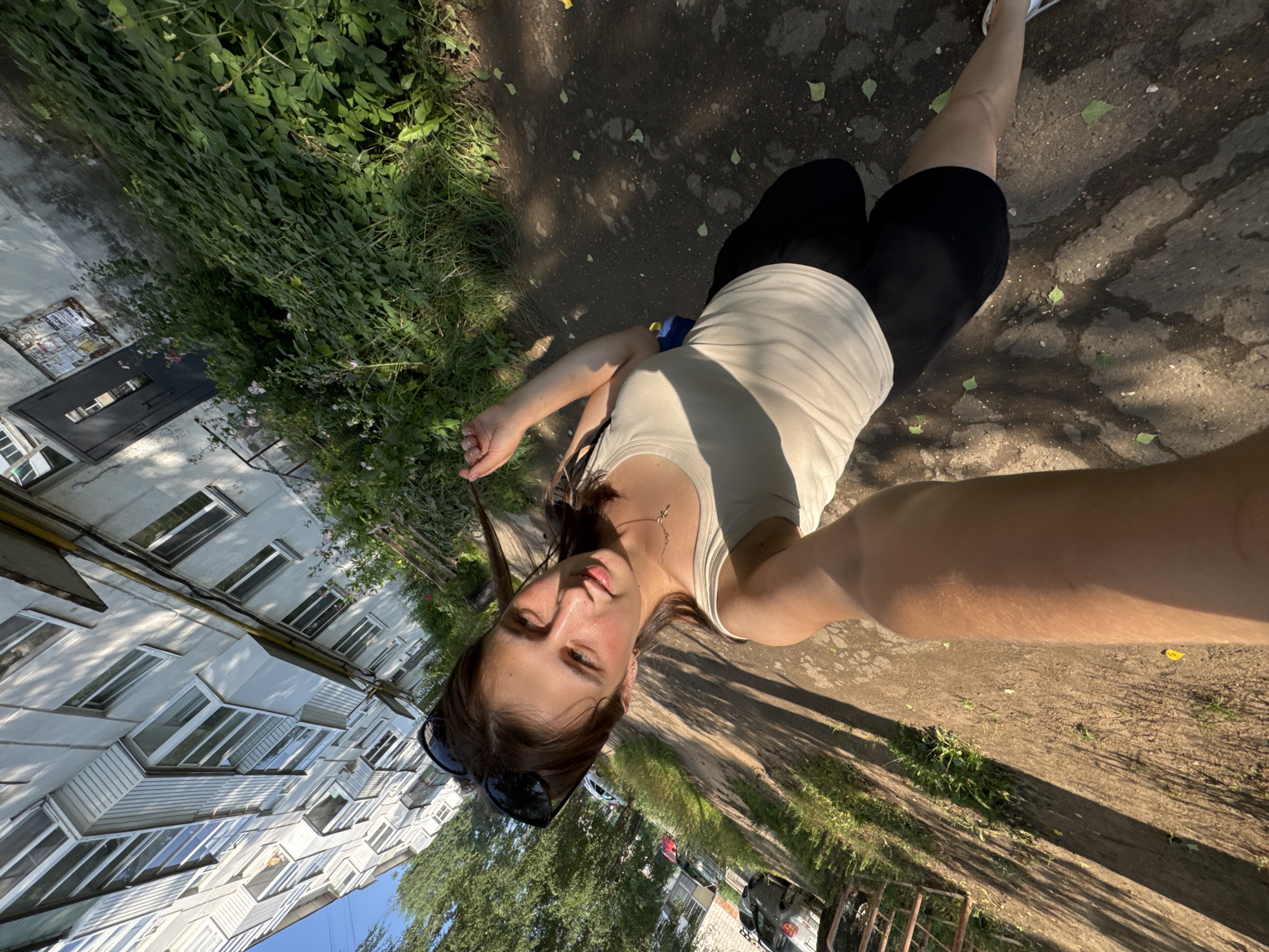 An outdoor, eye-level, wide-angle, full body selfie is captured by a young woman as she stands on a cracked, paved sidewalk in front of a multi-story apartment building in Chaikovsky, Russia. The woman has brown hair and wears sunglasses on her head, a cream-colored tank top, and black shorts. She is holding her phone to take the photo with her right arm outstretched. The sidewalk is lined with overgrown green bushes and trees, casting long shadows across the ground from the sunlight. The apartment building is light gray with numerous windows and balconies. Cars are parked along the street to the left of the frame.