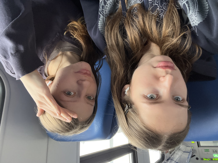 Two young females are seated inside a public transport vehicle, likely a train or intercity bus, situated in the city of Bryukhovetskaya, Russia. The interior features blue upholstered seats and a white overhead panel displaying "TEMP +7°C" and "01". The window background indicates an overcast exterior.
The female on the left has long, light brown hair, blue eyes, and light skin, and wears a dark hooded sweatshirt with a patterned design. A small, dark hair clip is visible in her hair above her right ear, and a white wireless earbud is in her right ear. She looks directly forward towards the camera with a neutral expression.
The female on the right has long, light brown hair, light-colored eyes, and light skin. She wears a dark blue V-neck sweater over a lighter-colored crew neck shirt. A white wireless earbud is in her left ear. Her right hand is raised, palm partially open, resting above her forehead. She is looking slightly towards the camera with a subtle smile. The image appears to be a close-up photograph or selfie, capturing both individuals from the chest up.