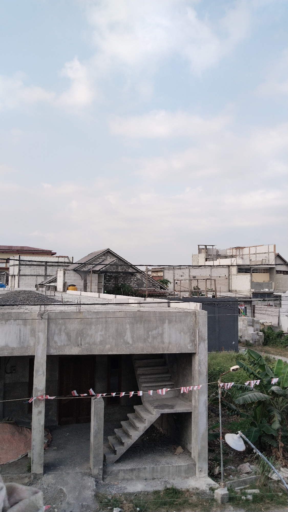 The image depicts a partly constructed concrete building in a semi-urban environment. The building features an open ground floor with a concrete staircase leading to an upper level. Small Indonesian flags are strung along a wire in front of the staircase. In the background, several other buildings are visible, some appearing to be under construction with exposed concrete and blockwork, while others are more complete. A pile of grey gravel is present on a concrete platform in the foreground. The sky is overcast with white clouds. The surrounding area shows some vegetation, including banana trees, and what appears to be a street lamp with two lights. The scene suggests ongoing construction or renovation activity in the city of Banjarsari, Indonesia.