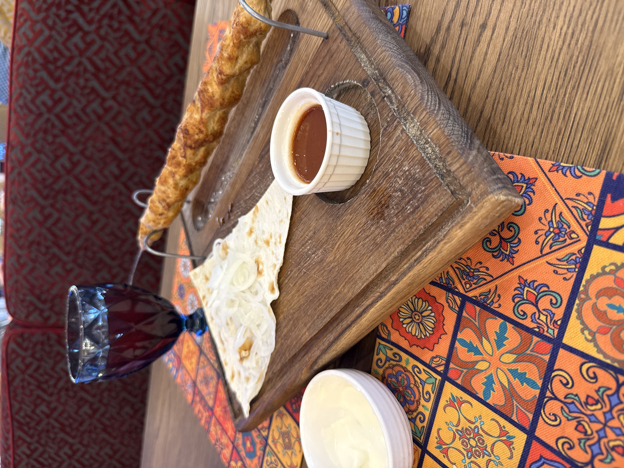 A close-up, eye-level shot shows a kebab skewer with a twisted appearance resting on a wooden serving board. Beside the kebab, a piece of flatbread is topped with thinly sliced onions. A small white ramekin holds a reddish-brown sauce, and another white bowl contains a creamy white dip. A blue glass filled with a dark red beverage is positioned to the left of the board. The entire arrangement sits on a table covered with a vibrant, colorful tablecloth featuring a geometric tile pattern. In the background, a red upholstered seat with a textured pattern is visible. The scene appears to be set in a restaurant in Saint Petersburg, Russia.