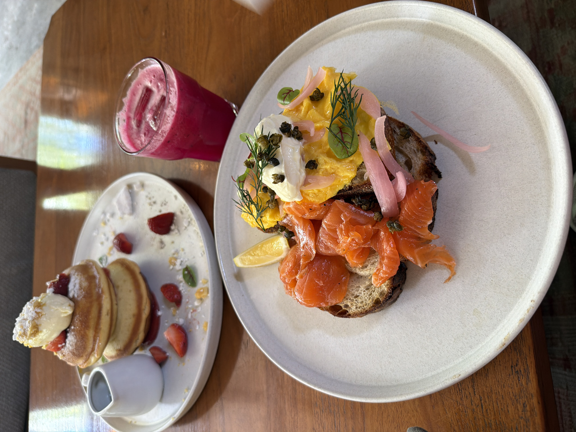 A high-angle shot depicts two plates of food and a drink on a wooden table. The plate on the right contains what appears to be a breakfast dish with smoked salmon, scrambled eggs, pickled onions, dill, and capers, served on top of a slice of dark bread. A lemon wedge is also on the plate. The plate on the left contains pancakes with what looks like cream or whipped butter on top, with a small carafe of syrup. There are also cut strawberries and what might be basil leaves decorating the plate. Between the plates is a glass of a pink-colored beverage with ice cubes. The setting seems to be a restaurant. The city of Unknown, Unknown cannot be confirmed based on the image.