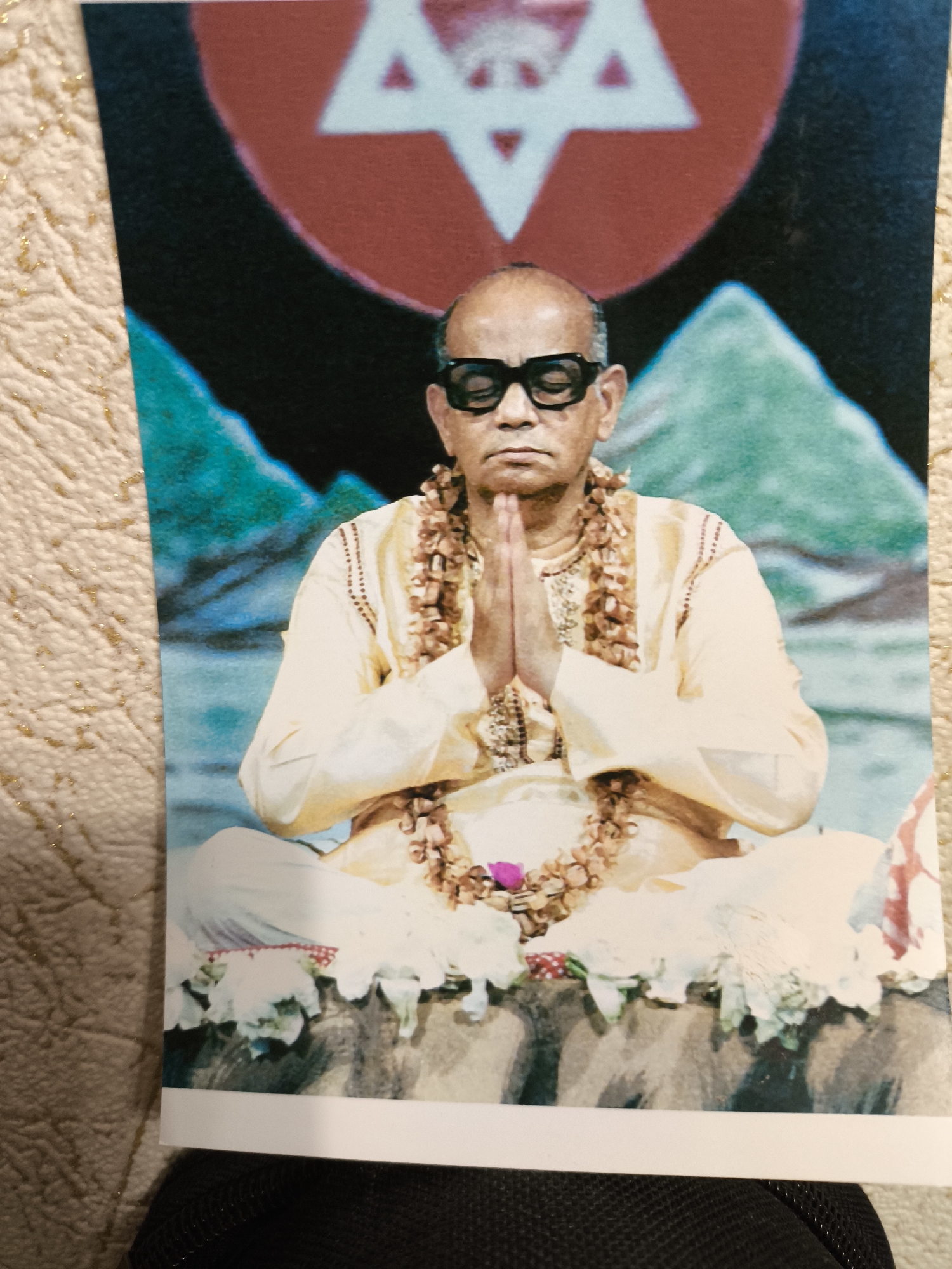 A photograph shows an Indian guru or spiritual leader sitting in a meditative pose with hands in prayer. He is wearing eyeglasses and a light-colored robe adorned with garlands. Behind him is a backdrop with a red circle containing a white star, with blue and green landscape elements resembling mountains. The scene appears to be indoors or on a stage. The location cannot be determined from the image; Moscow, Russia is not evident.