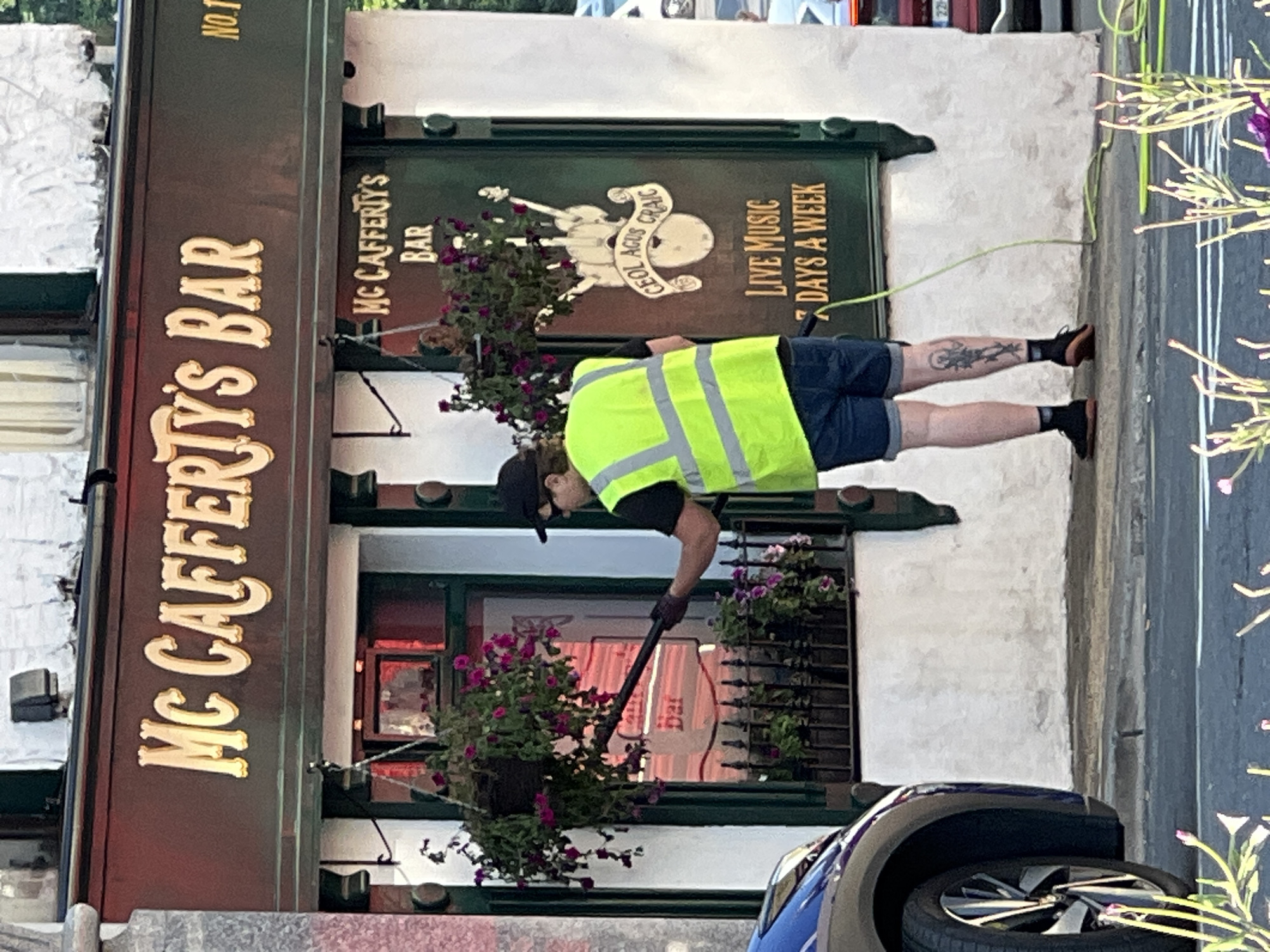 The image depicts the exterior of "Mc Cafferty's Bar" located in Donegal, Ireland. A person wearing a high-visibility vest and a baseball cap is seen from the side, holding a cleaning tool extended towards hanging baskets of flowers. The bar has a dark-colored facade with prominent gold lettering spelling out "Mc Cafferty's Bar". Below this, a green door or window section displays the text "Mc CAFFERTYS BAR" and "LIVE MUSIC 7 DAYS A WEEK", along with a graphic of a harp. The scene is set against a white stucco building. A blue car is partially visible in the foreground on the left. The street and sidewalk area in front of the bar are also captured in the image, with some greenery in the lower right corner.
