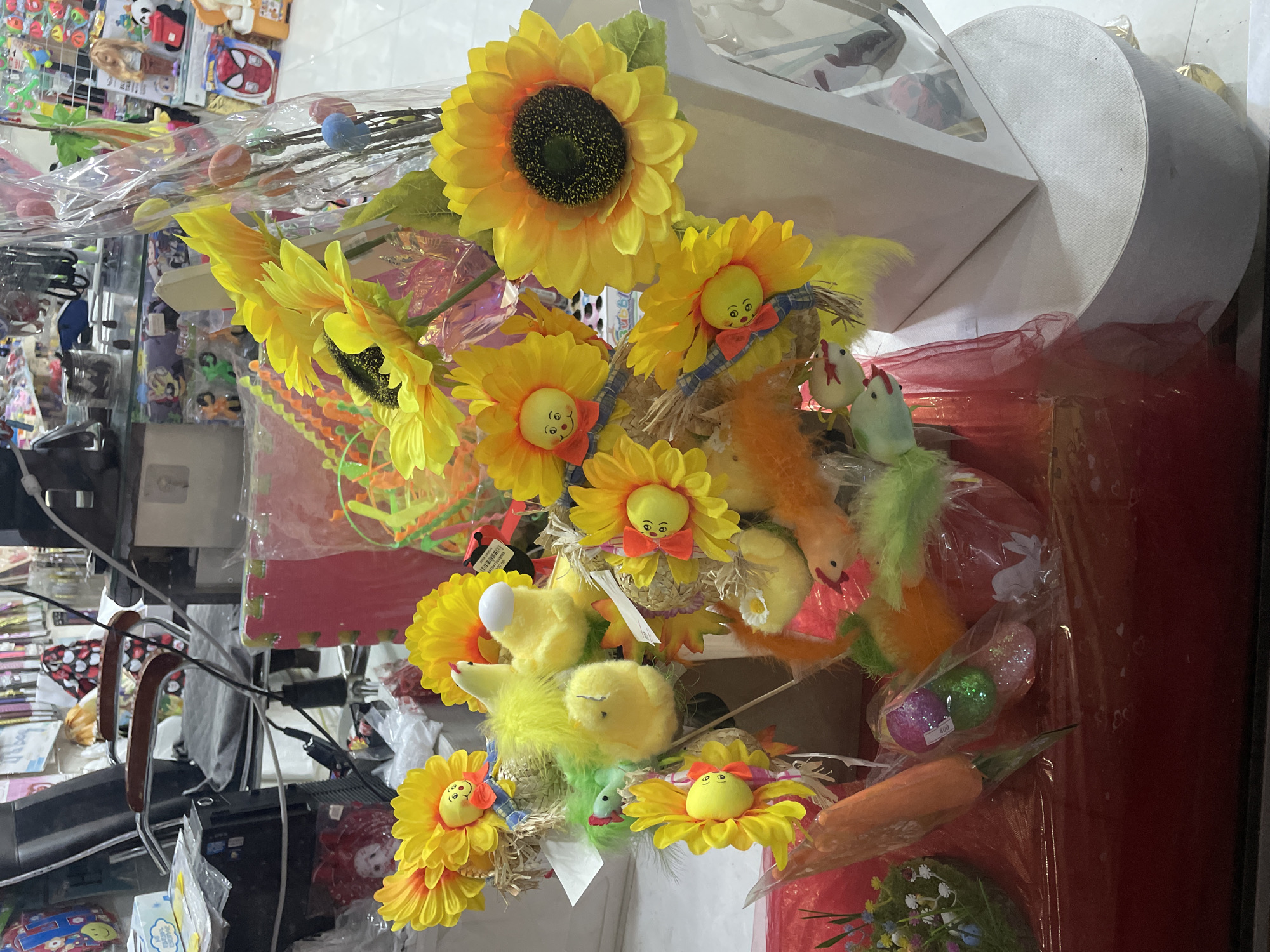 The image depicts a chaotic scene indoors, possibly within a shop in Yerevan, Armenia. The foreground is dominated by an Easter display, featuring artificial sunflowers, some with anthropomorphic faces and bow ties. Plush yellow chicks are also present, alongside decorative eggs and carrots, all seemingly part of an Easter arrangement. The arrangement is housed in a clear packaging, which obscures some details behind it.
Behind the Easter display, the environment appears cluttered. Shelves stocked with miscellaneous items are visible, including what seems to be toys and other packaged goods. There is also a chair and other furniture partially visible, contributing to the crowded and disorganized atmosphere. The lighting is bright and somewhat uneven, further emphasizing the busy nature of the scene.