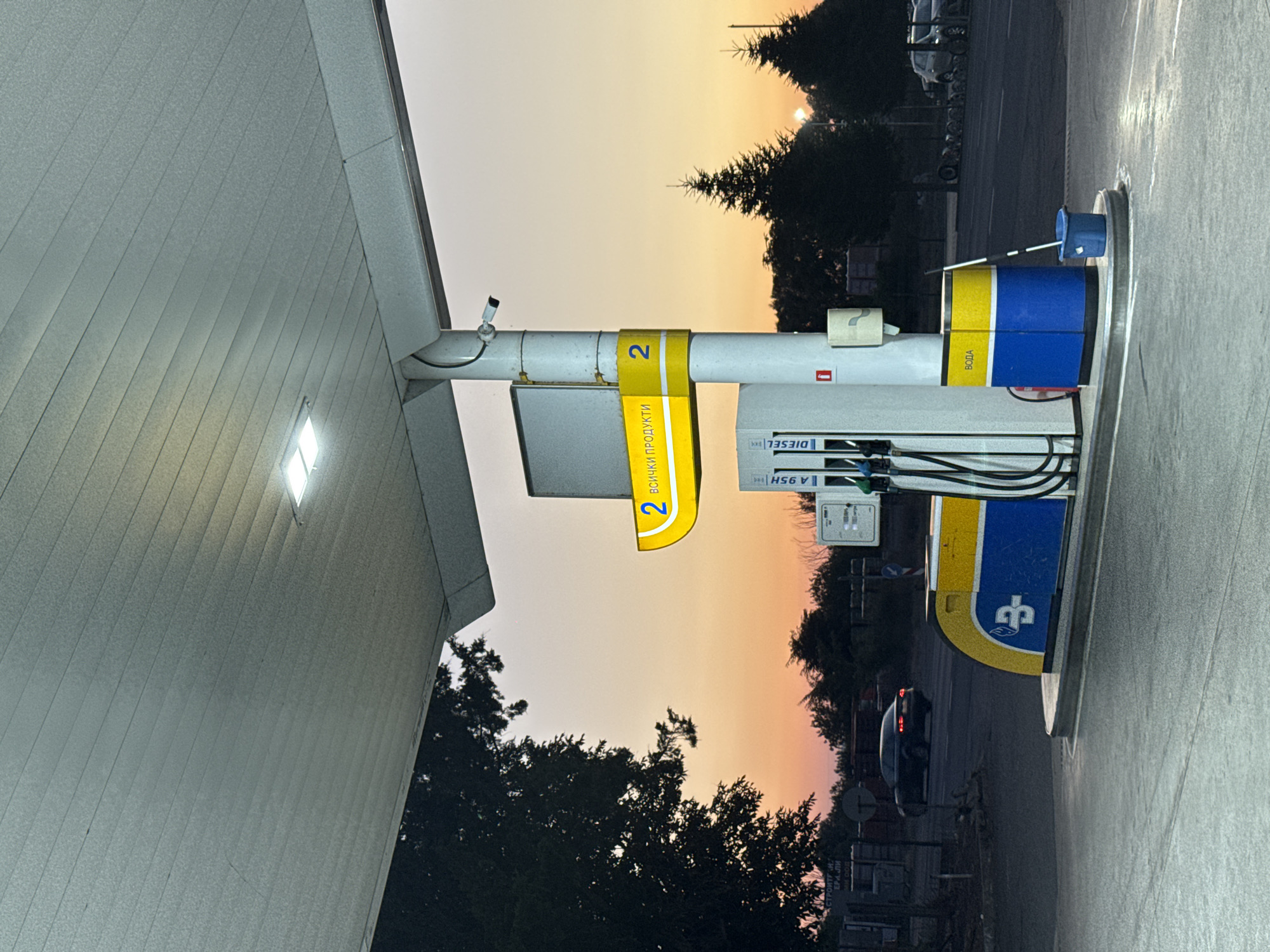 The image depicts a gas station in Sredets, Bulgaria, at dusk. A gas pump with yellow and blue branding stands prominently in the foreground, with a bucket and mop to its right. Above the pump, a yellow sign displays the number "2" and text in Bulgarian. A security camera is mounted on the central pillar supporting the station's canopy. To the left, silhouetted trees frame a dusky sky with hues of orange and pink. In the background, a dark-colored car drives away, its taillights illuminated. Further back, more trees and parked vehicles are visible. The overall scene suggests a quiet, possibly end-of-day atmosphere at a roadside service station.