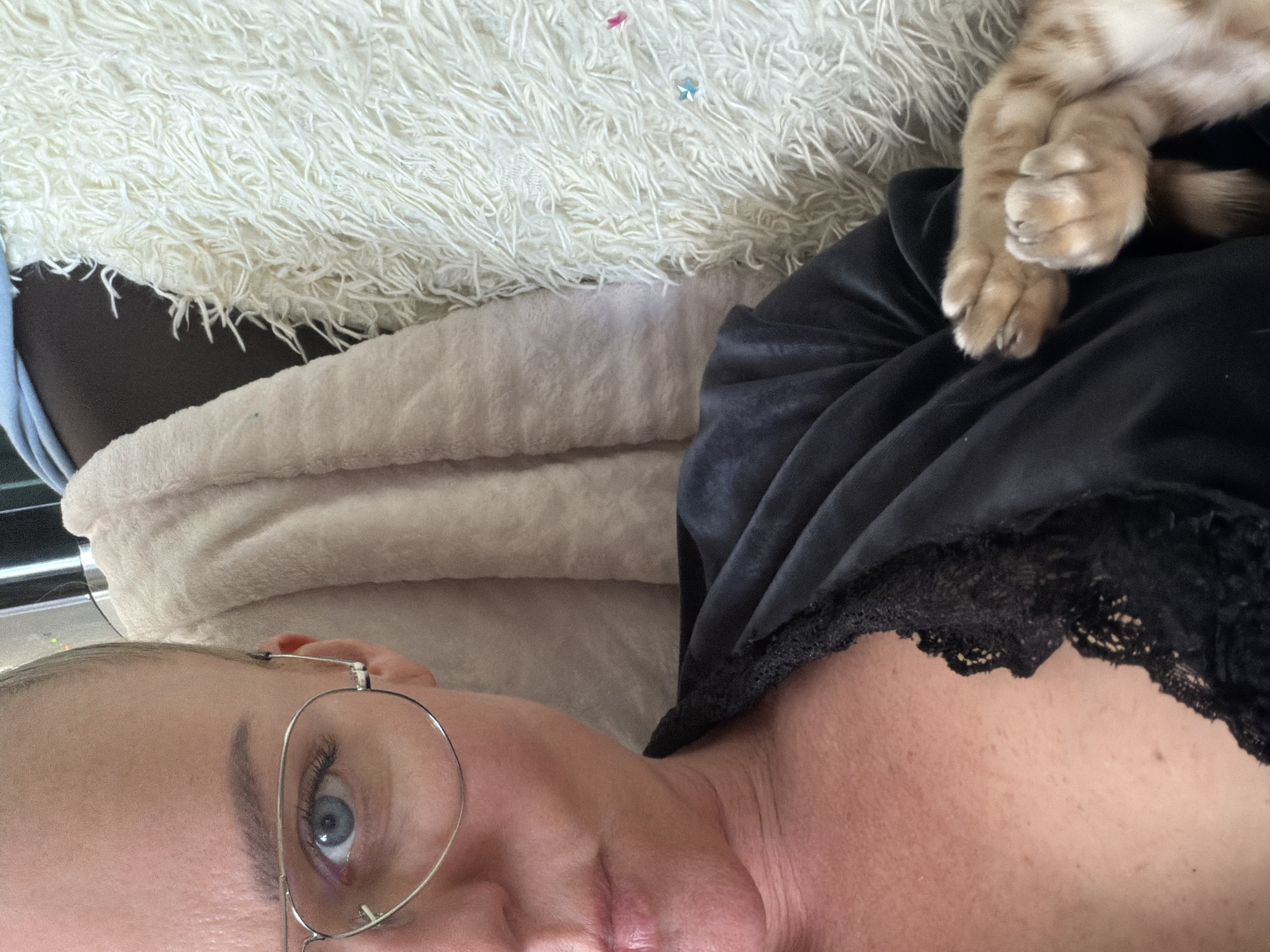 A low-angle, close-up shot depicts a person, likely female, lying down. Part of their face, specifically the eye with silver-rimmed glasses, nose, and part of the mouth, is visible. They have fair skin. The person is wearing a black, lace-trimmed garment. A cat's paws rest on the garment. The background consists of a fluffy white blanket and a tan, possibly folded blanket or pillow. There is no indication of a specific location in Vsevolozhsk, Russia, or any other location.