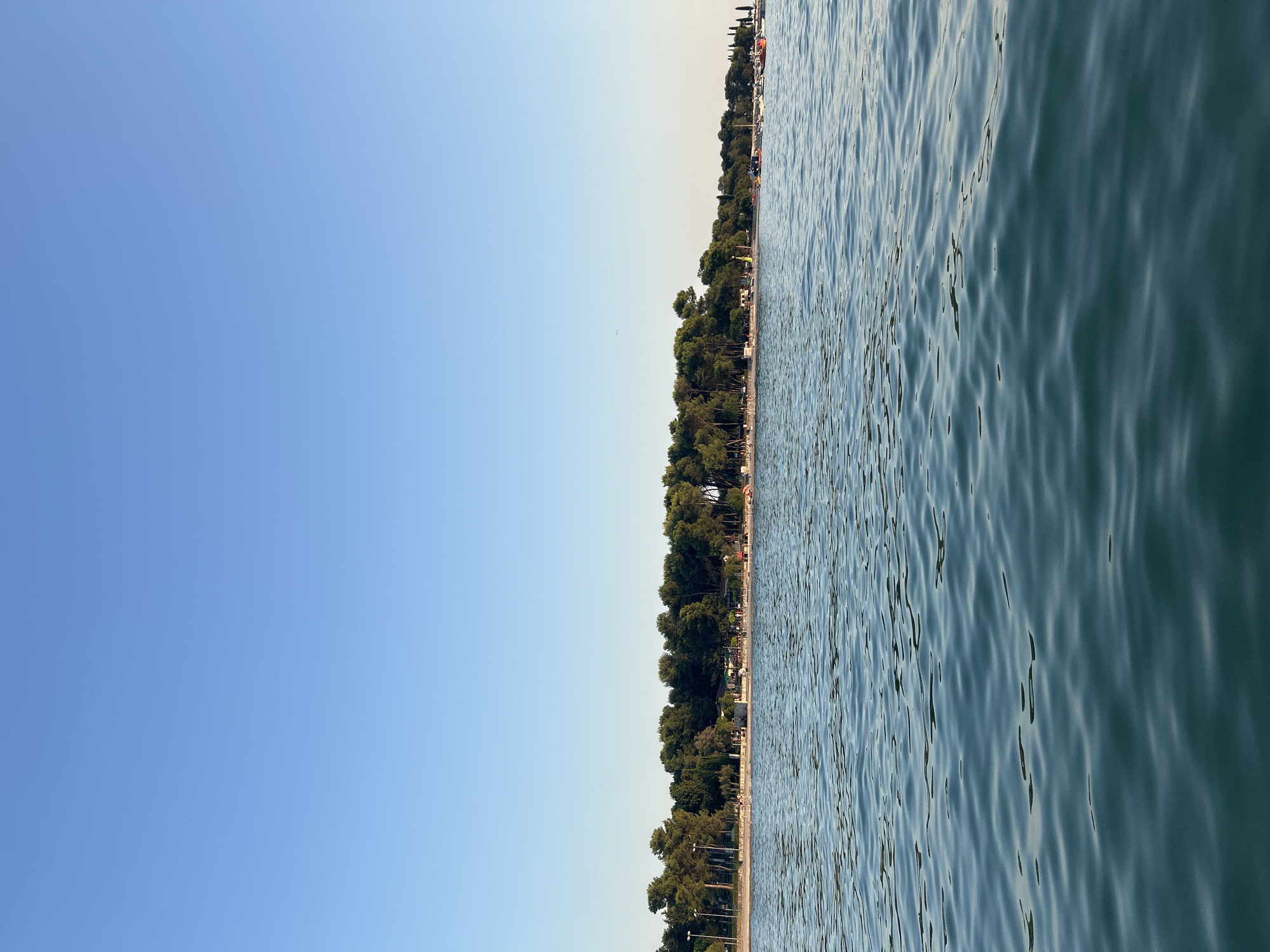 The image depicts a tranquil scene of the Adriatic Sea in Katoro, Croatia. The foreground is dominated by the rippling surface of the turquoise water, reflecting the clear blue sky above. In the mid-ground, a sandy beach is lined with a dense row of pine trees. Several structures, possibly beachside cafes or amenities, are visible nestled among the trees. Small groups of people are scattered along the beach, suggesting a leisurely afternoon. In the distance, further along the coastline, more trees and some boats moored in the water can be observed. The overall impression is one of a peaceful summer day at a coastal resort.