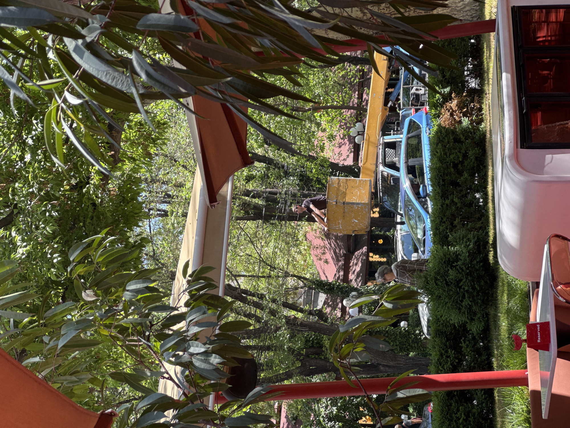 The image depicts an outdoor scene, likely a restaurant or cafe, in Yerevan, Armenia. In the foreground, foliage and branches frame the view, while red structures and a white booth with a "Reserved" sign are present. Further back, individuals are working on a crane or truck, visible in the middle ground among trees and greenery. There are various plants, adding to the outdoor setting. The background shows buildings and possibly a street, completing the urban backdrop.