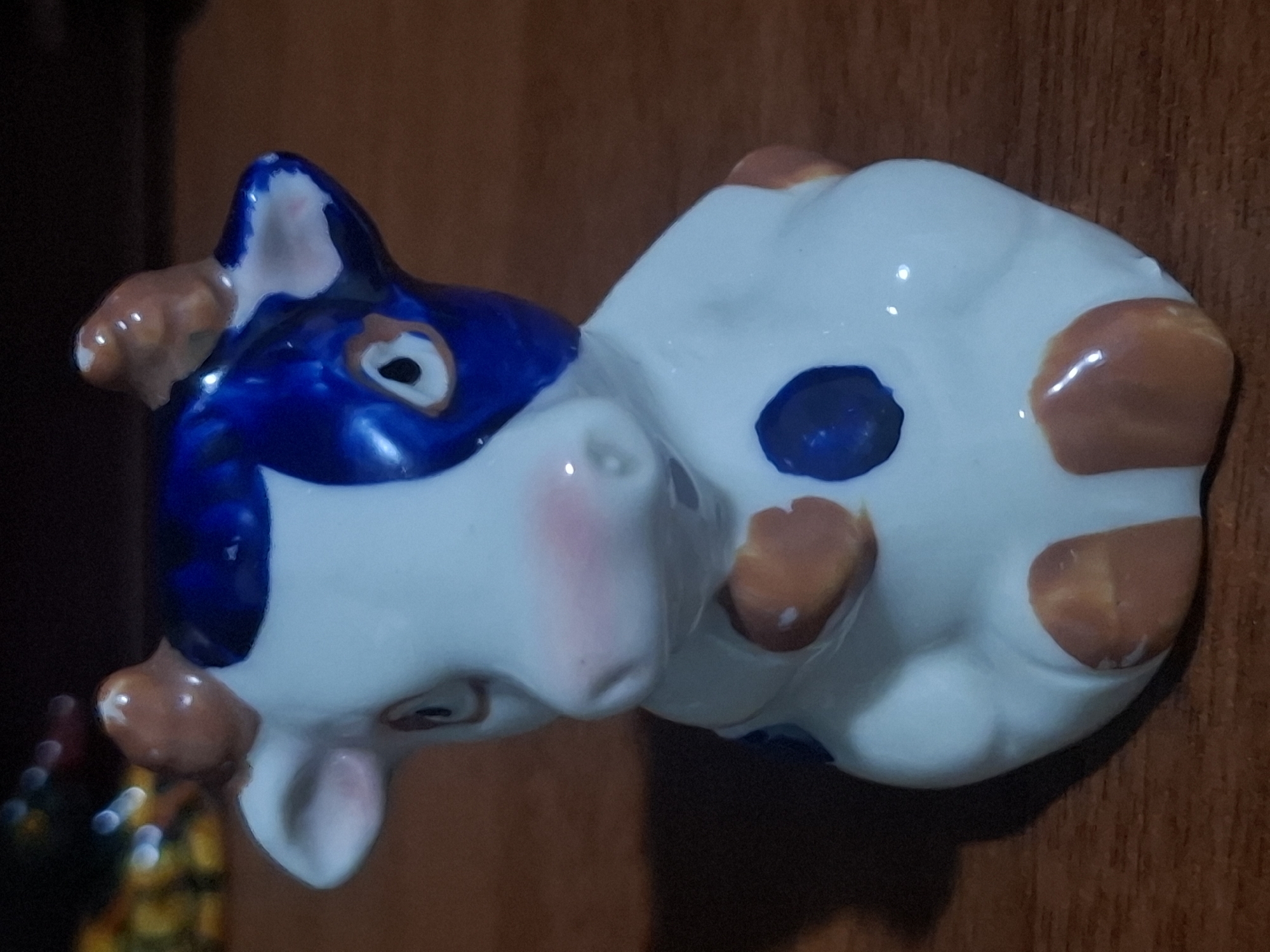 A ceramic figurine of a cow is positioned on a wooden surface, likely a table. The cow is white with blue patches on its head and body, and brown patches on its ears, horns, and legs. It has pink accents on its nose and cheeks, and black eyes. The background is dark and out of focus, showing some blurred colorful objects to the left and a dark shadow to the left of the figurine. The lighting appears to be artificial, creating reflections on the glossy surface of the figurine. The exact location within Almaty, Kazakhstan, cannot be determined from the provided image.
