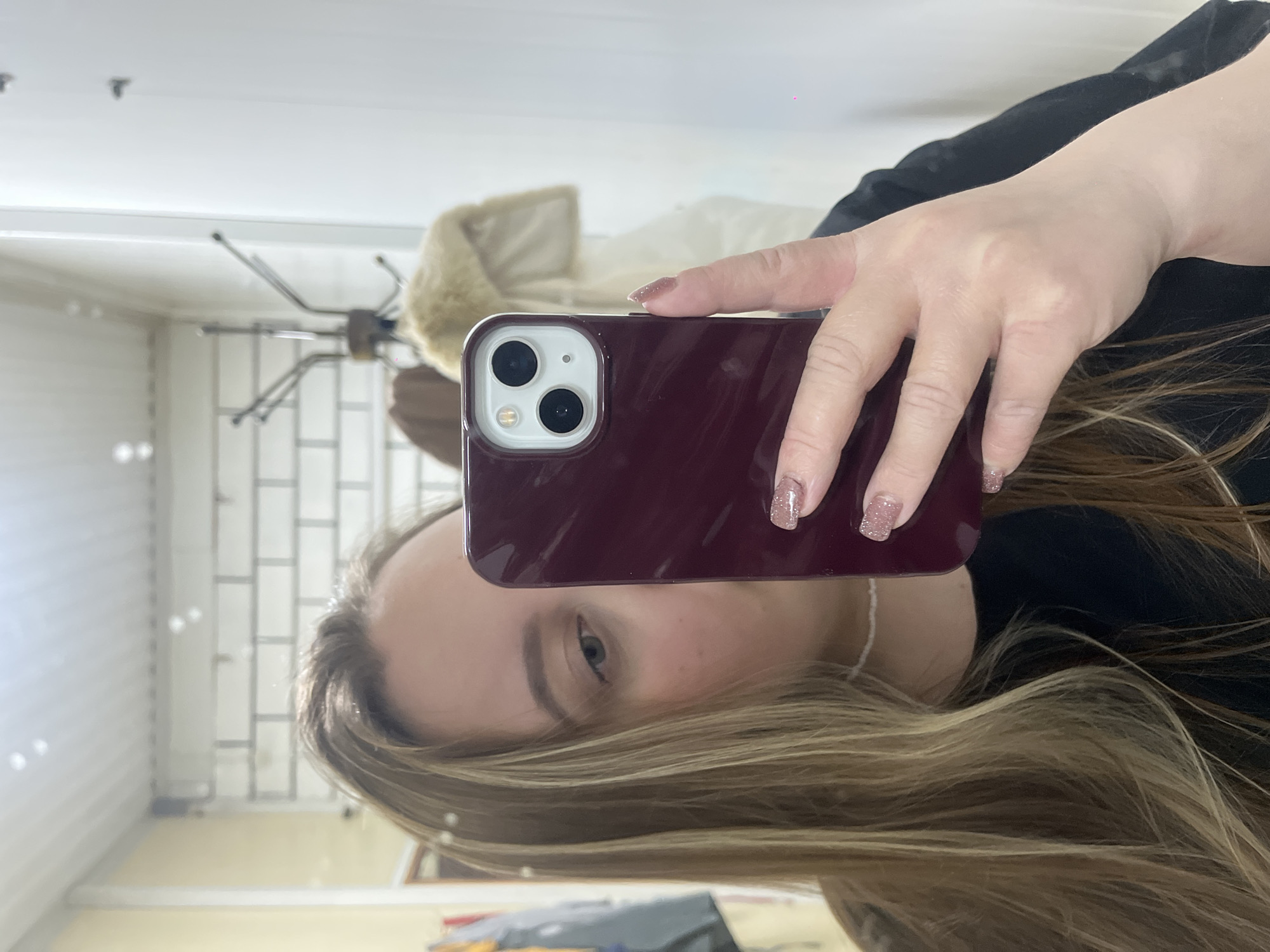 A person is taking a selfie in a mirror. The person is holding a smartphone with a burgundy case. Their right hand is visible holding the phone, and their face, hair, and a portion of their upper body are visible. The person has blonde hair with dark roots, fair skin, and painted fingernails. A white coat hangs on a coat rack in the background. The background also shows a door or wall with white paneling, and a glimpse of a cluttered room. There is no information in the image to indicate this photo was taken in Novopokrovskaya, Russia.