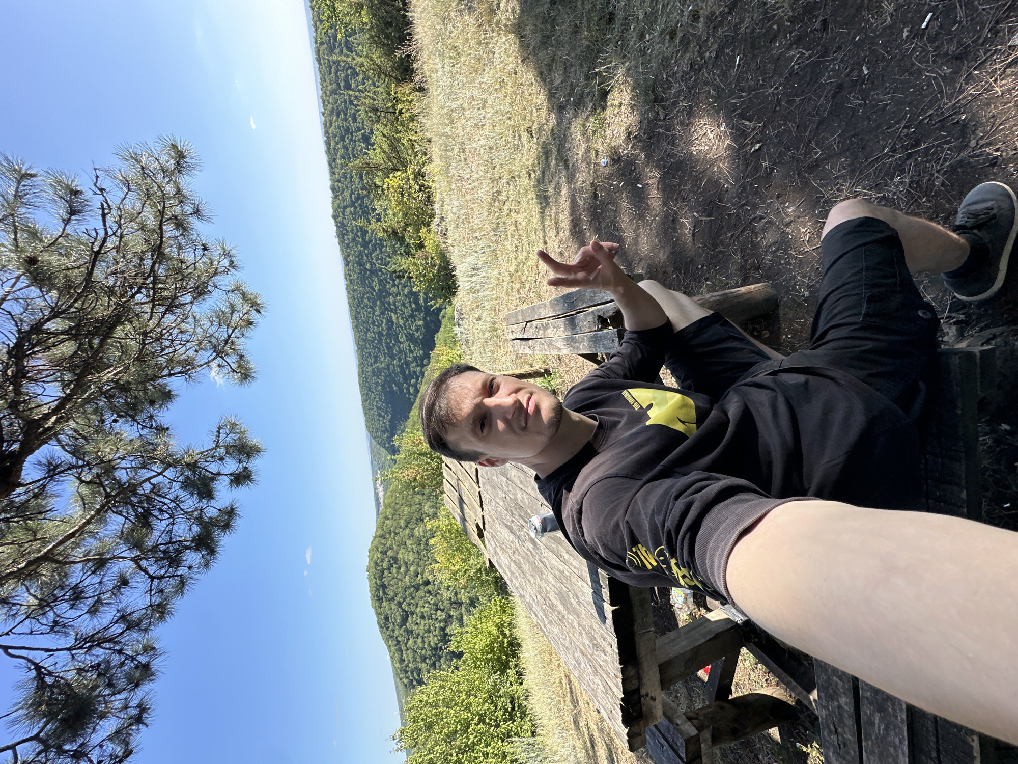 A man is seated on a wooden bench at a picnic table, taking a selfie. He is wearing a black hoodie with a yellow emblem and black shorts. The man is looking at the camera and making a peace sign with his right hand. Behind him, there is a scenic view of rolling green hills and a clear blue sky. The picnic table appears weathered and aged. The surrounding ground is covered with dry grass, dirt, and some scattered debris. The location is identified as being in the city of Nizhyn, Ukraine, likely an outdoor recreation area or viewpoint.