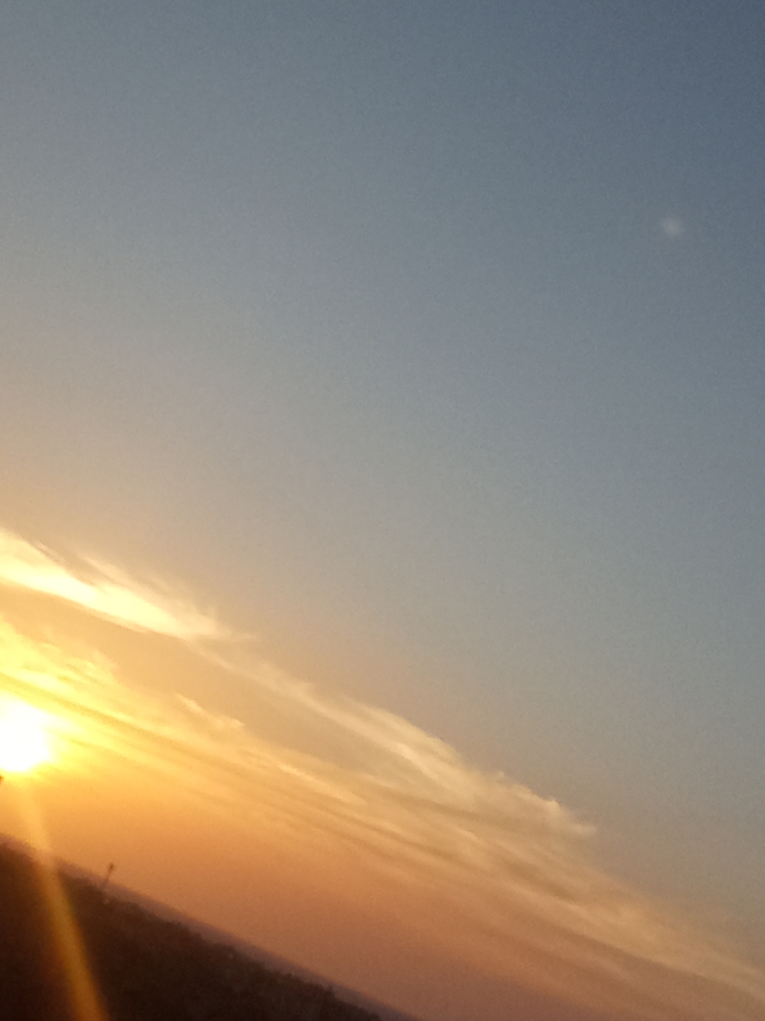 The image shows a sky at sunset in Juan Griego, Venezuela. The sun is low on the horizon, casting an orange and yellow glow on the clouds. The sky is mostly blue. In the lower part of the image is a dark silhouette of land.