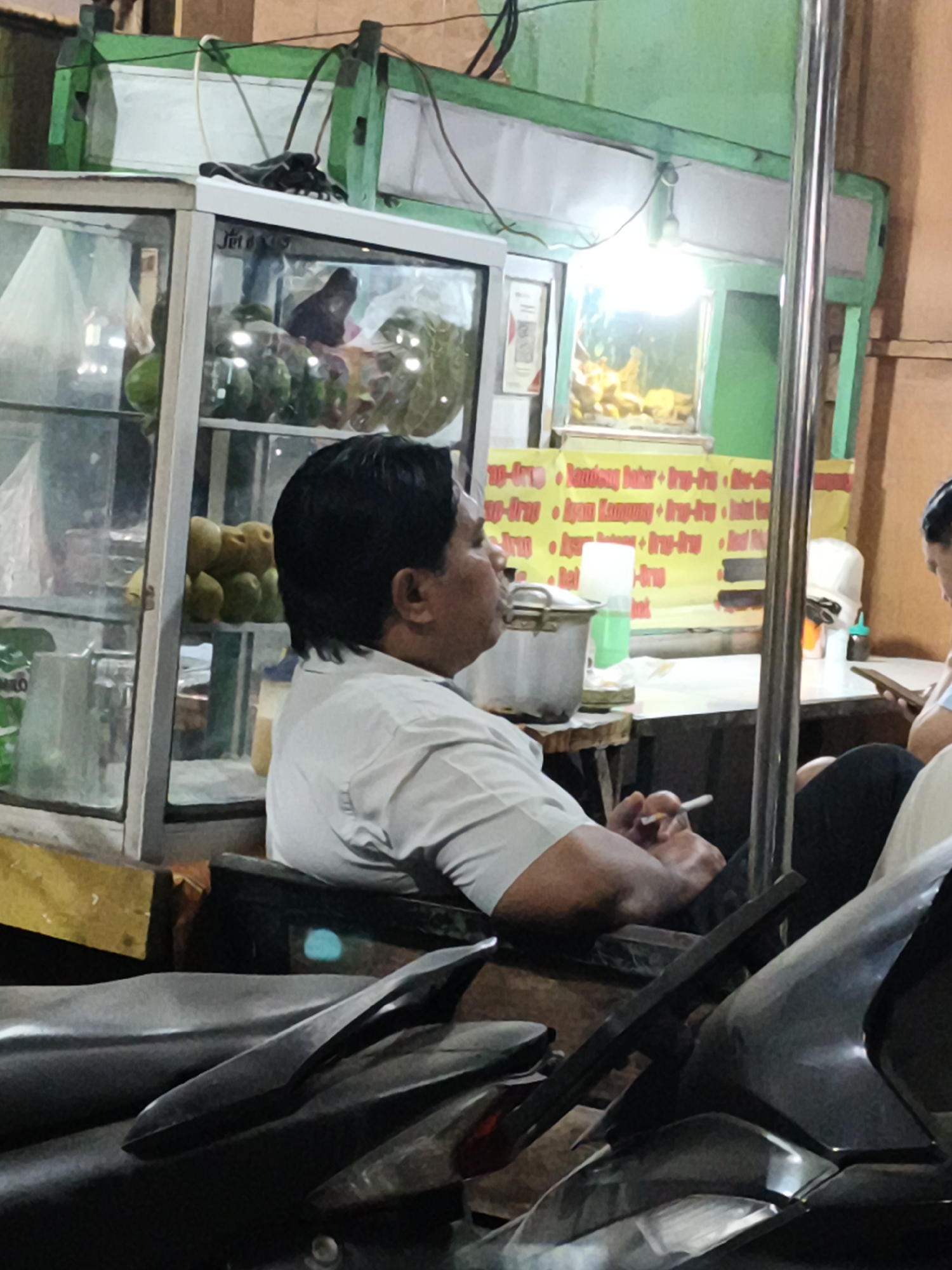The image shows a street food stall in Petiken, Indonesia. In the foreground, there are parts of two motorcycles. A man wearing a light-colored shirt is seated and smoking a cigarette. Behind him, a display case contains various items, possibly food, including some green fruits and yellow fruits. A metal pot and a green bottle are visible on a wooden stool. Further back, there is a yellow banner with Indonesian text. The stall has a green structure overhead with lights. Another person, partially visible, is seated to the right, holding a book or tablet. The overall scene appears to be at night or in low light conditions.
