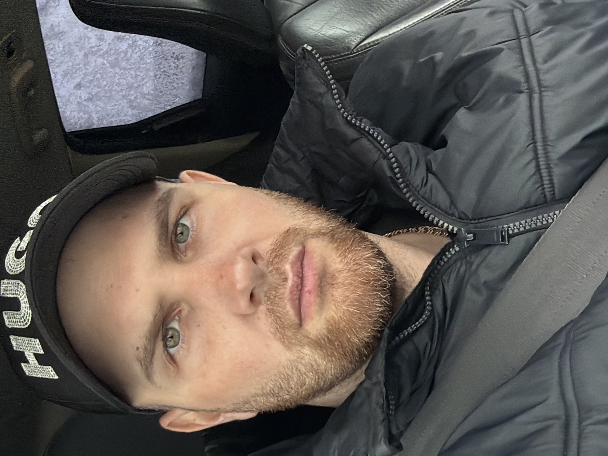 A selfie of a man with a light complexion and a reddish-brown beard, wearing a black baseball cap with white lettering, taken inside a vehicle. He has green eyes and is wearing a black puffer jacket and a seatbelt. The interior of the vehicle appears to be black leather. The background, visible through the car window, is out of focus and appears to be a gray or white surface. The location in the city of Chaikovsky, Russia is not ascertainable from the image.